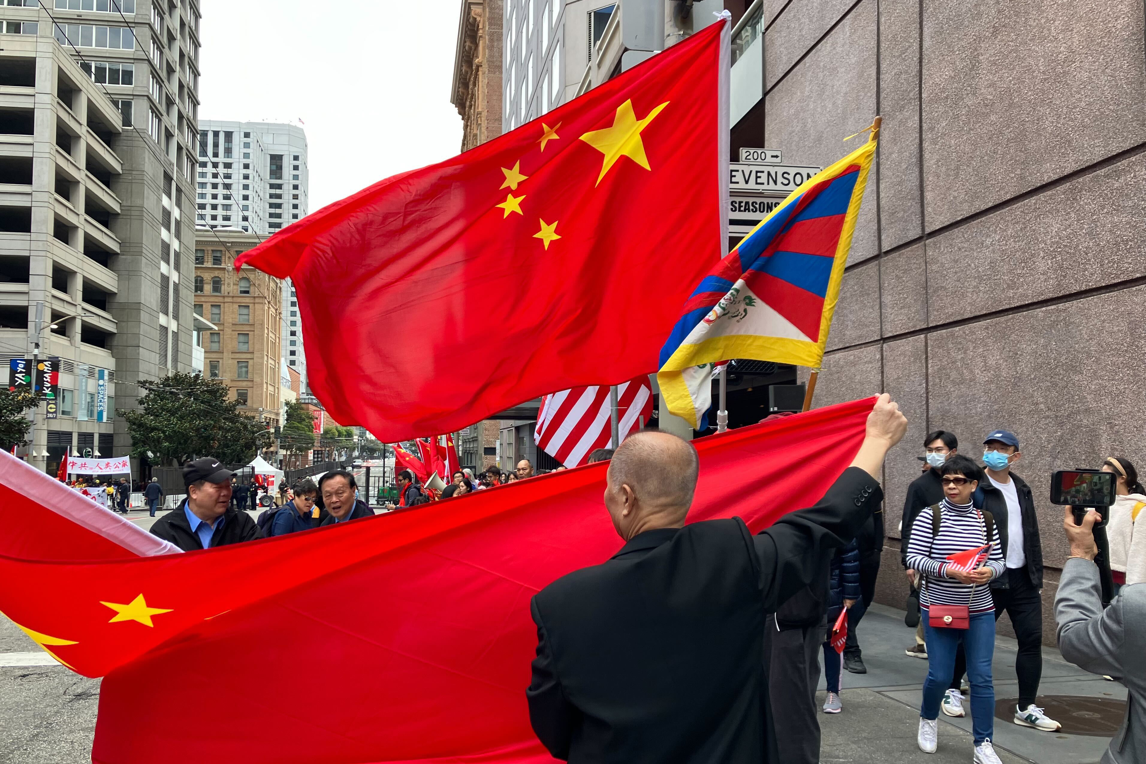 A man stretches a large, red, Chinese flag around a Tibetan flag on a city sidewalk as pedestrian pass.