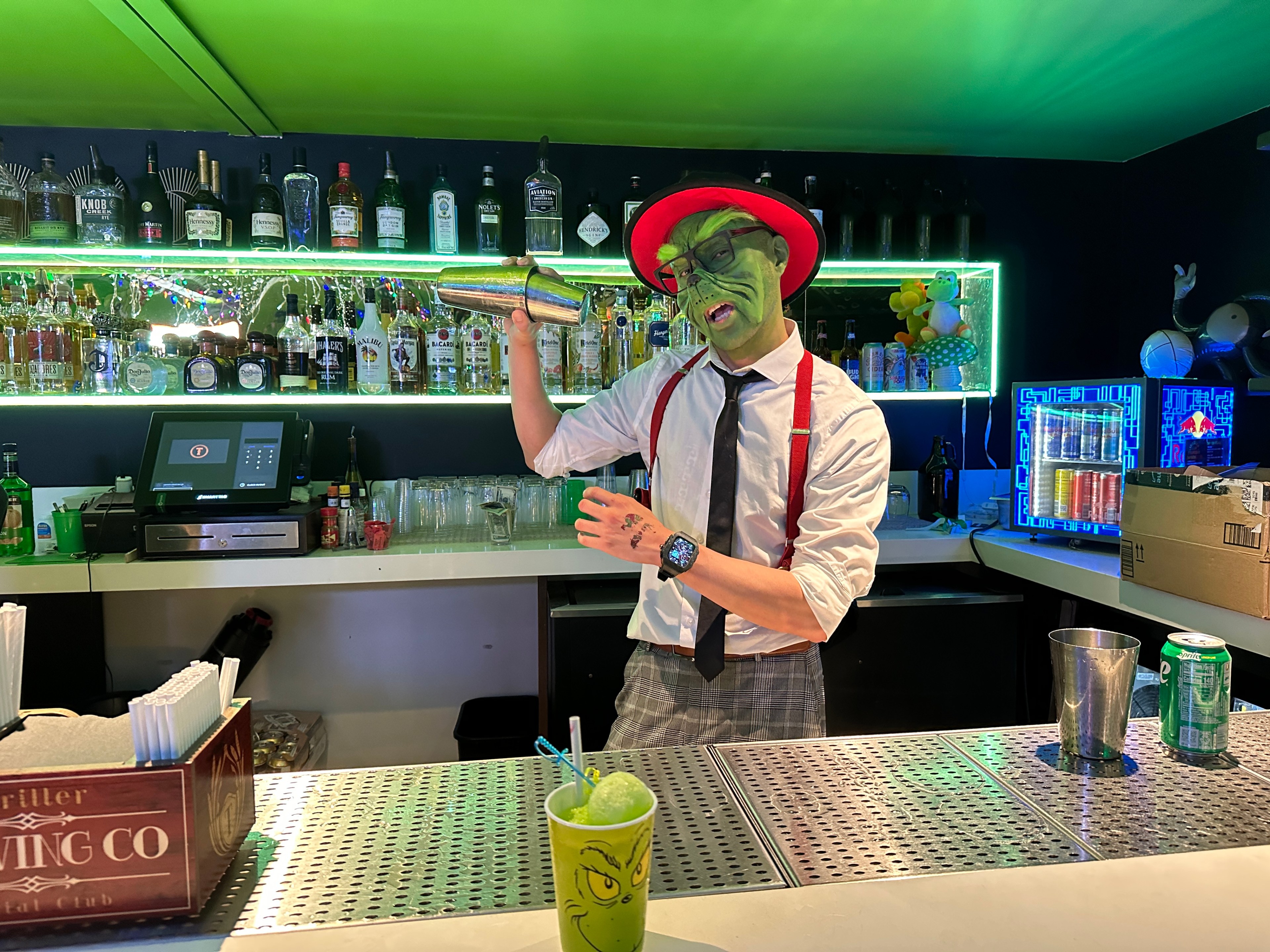 A bartender dressed as Dr. Seuss's the Grinch shakes up a Grinch-themed green cocktail behind a bar.  