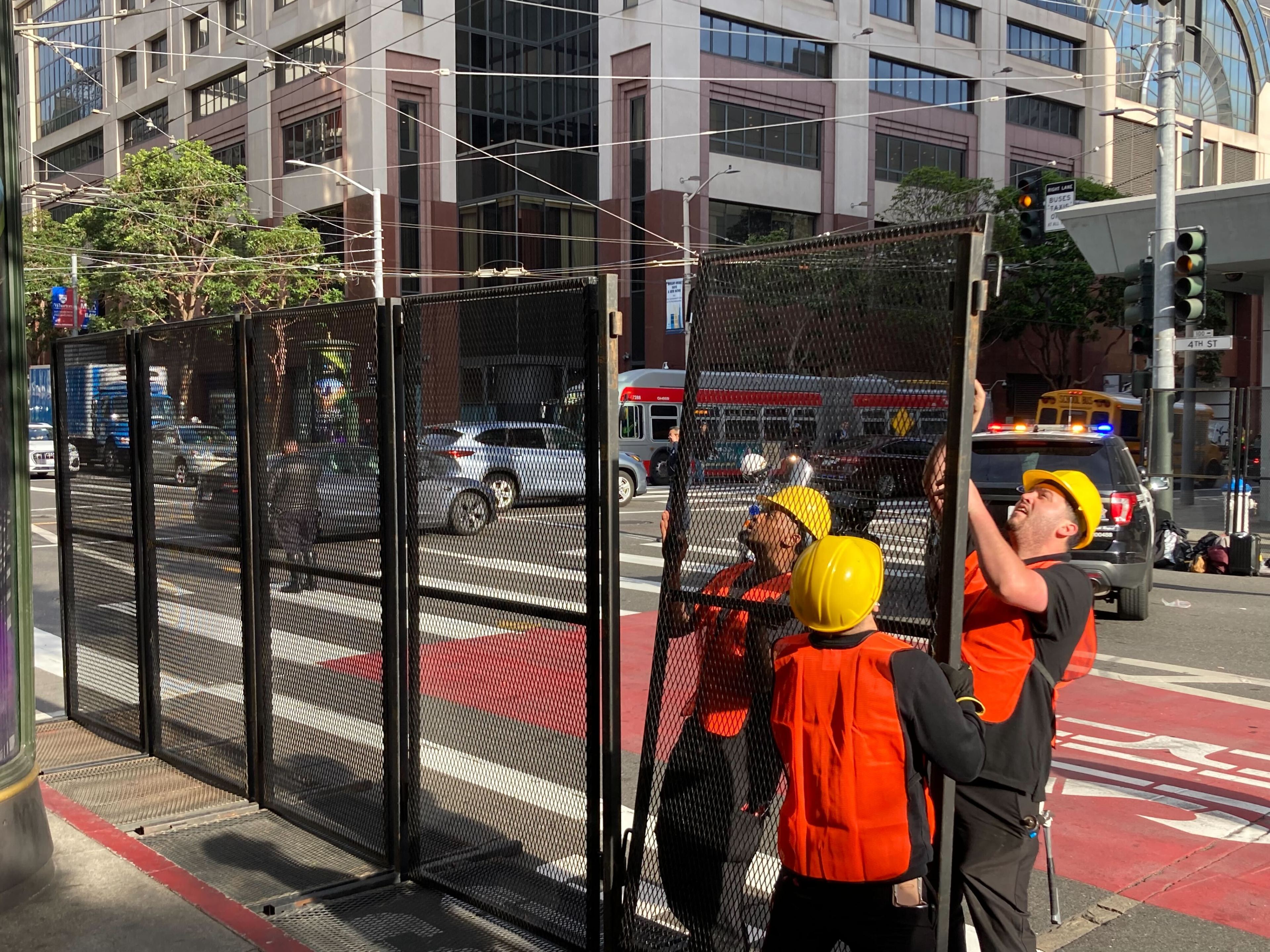 A group of construction workers putting up fencing on a public street with a police vehicle, cars and a bus in the background.