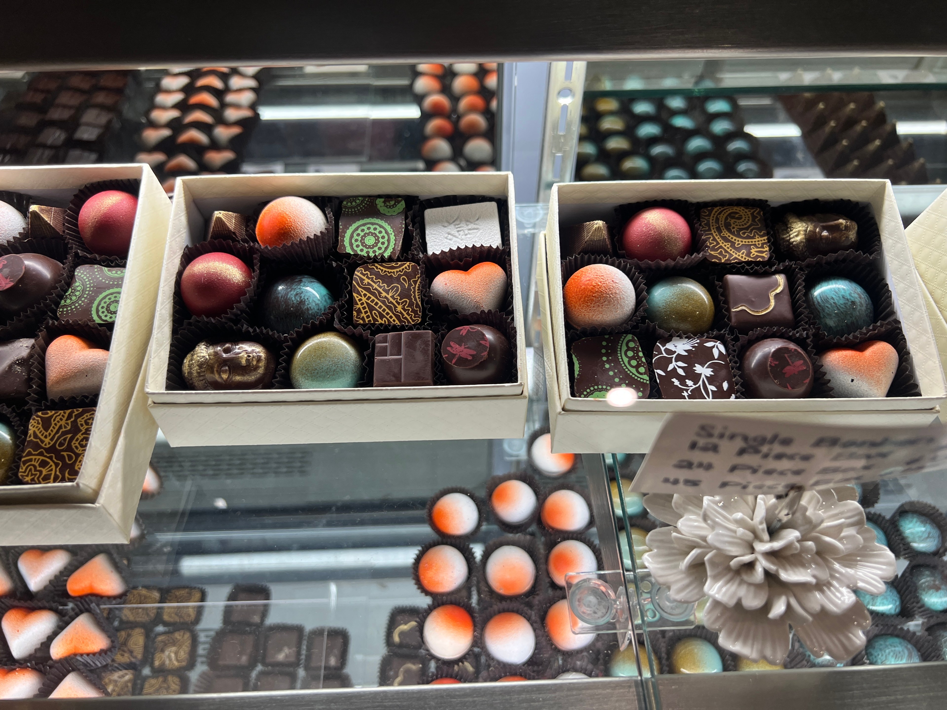 Pieces of chocolates in colors ranging from orange to blue and with different patterns are visible through a glass display case.