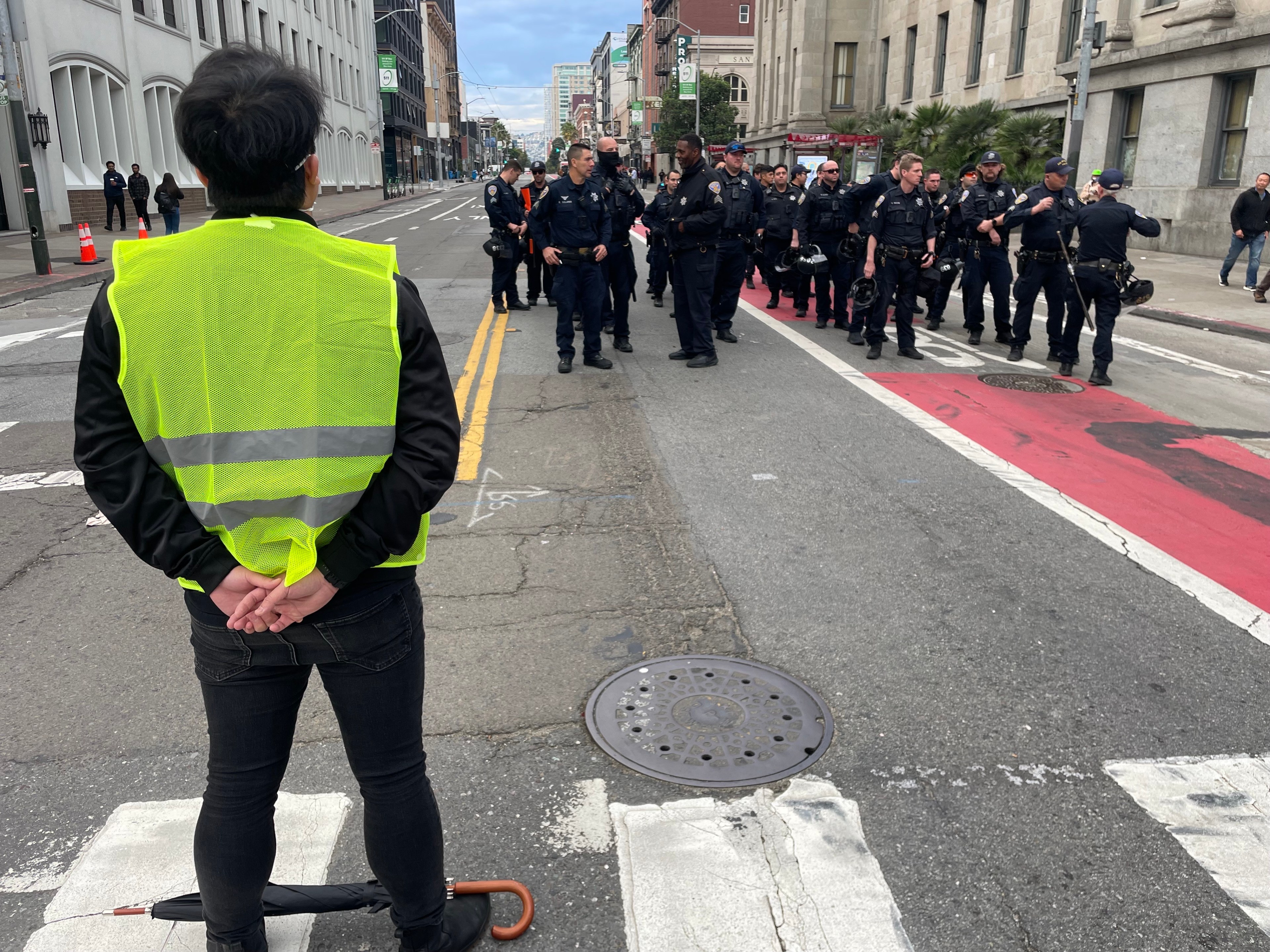 A person wearing dark clothing with arms folded behind them stands in a crosswalk a short distance from a group of police in dark colored uniforms in the middle of a street.