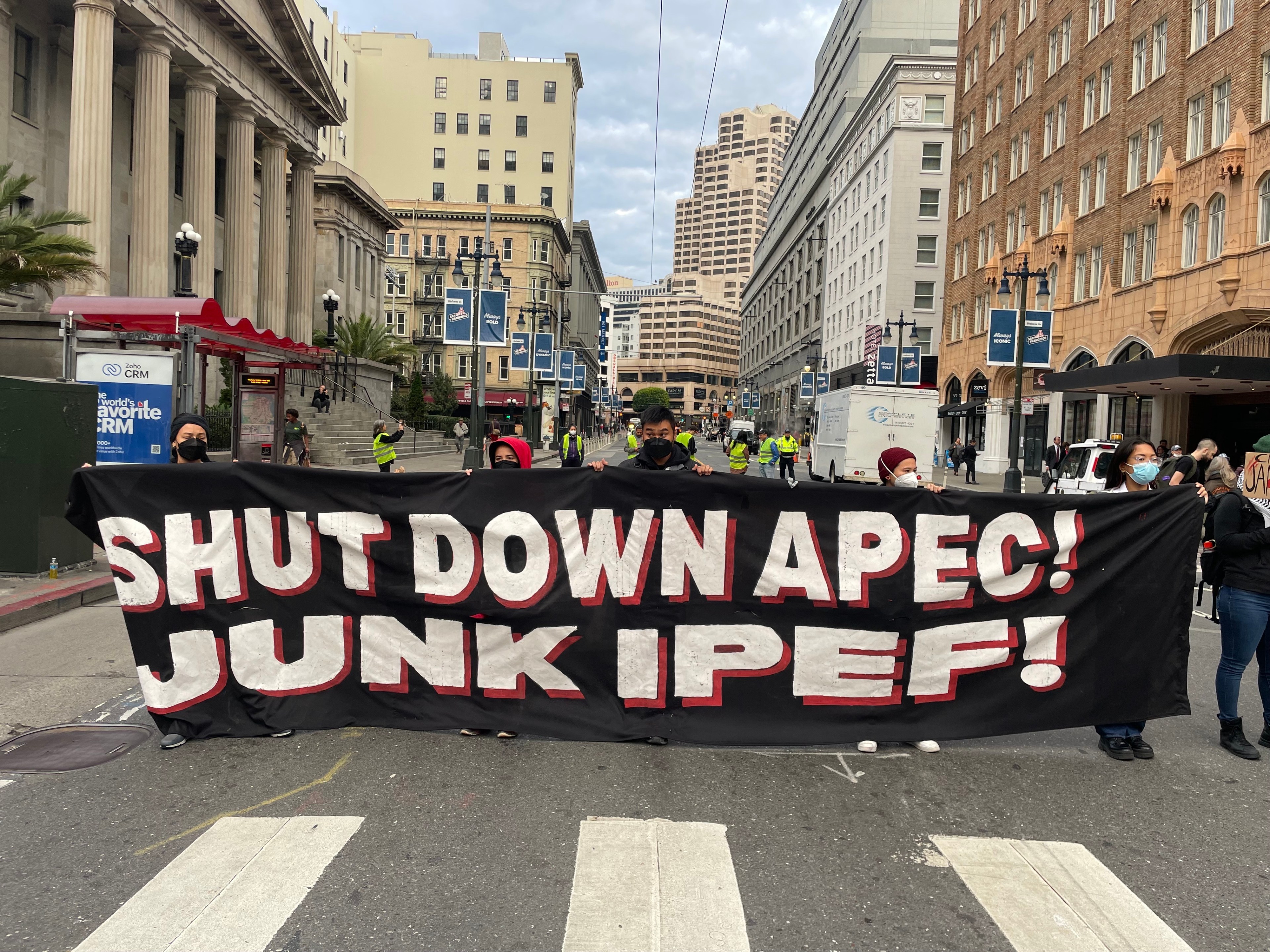 Several mask wearing protesters hold a dark colored banner with the words &quot;Shut down APEC! Junk IPEF&quot; across it in white block lettering while standing in a street blocked off to traffic.