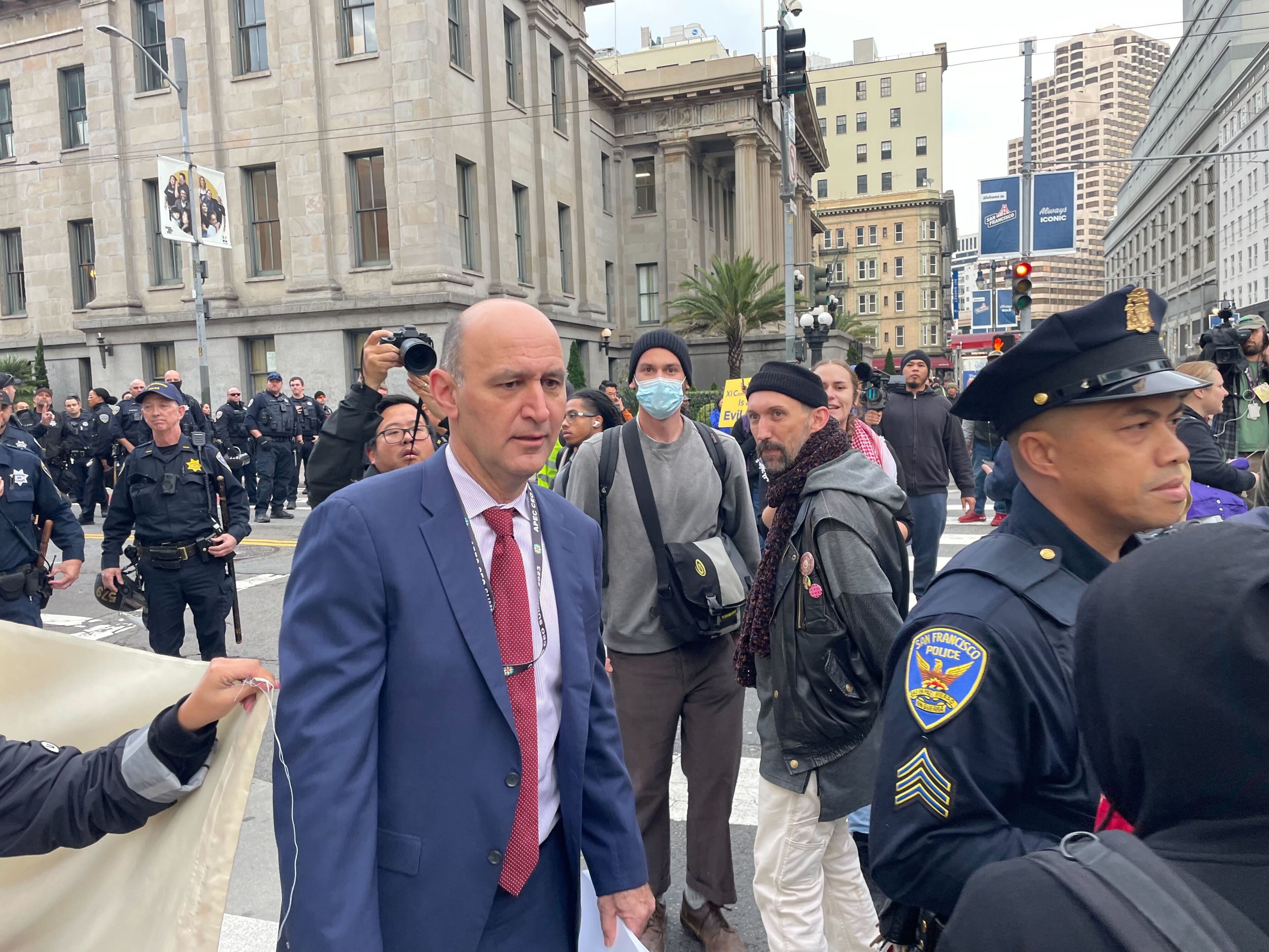 A shaven-headed suit-wearing man threads his way past uniformed police officers and a few protesters on a street corner.