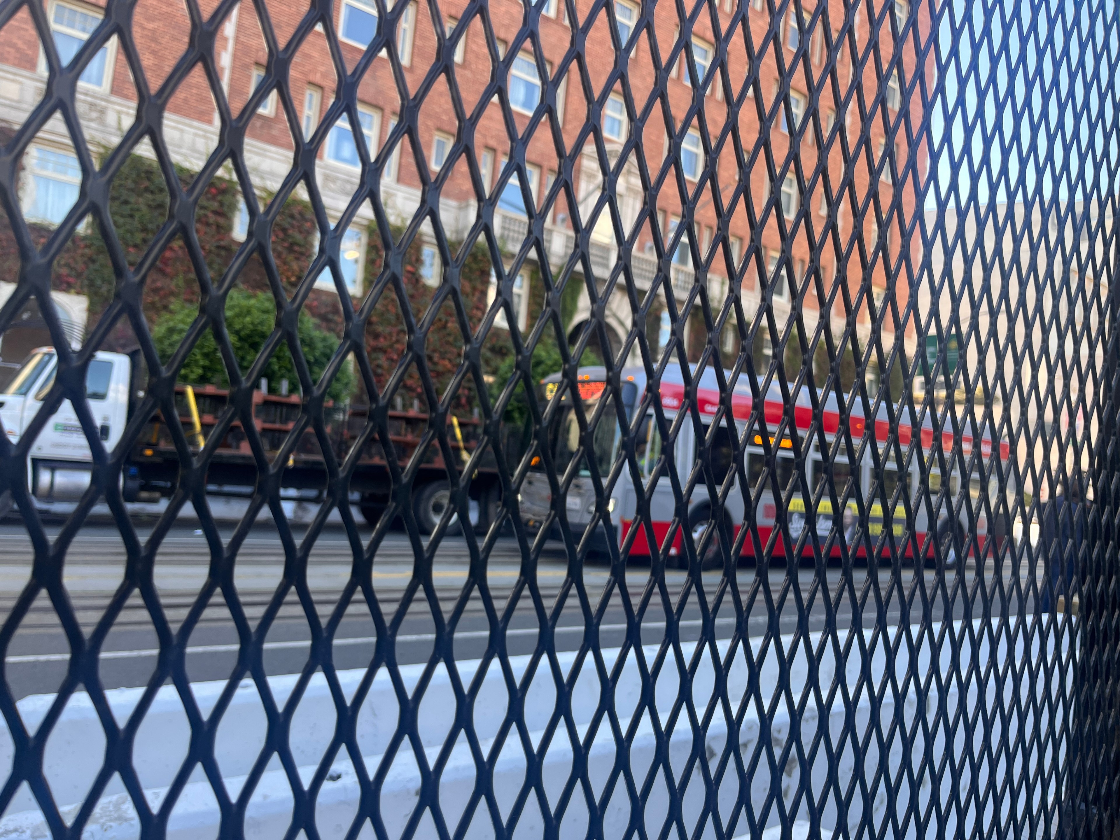A red bus is seen traveling down a street; the view of the bus is partially blocked by a security fence. 