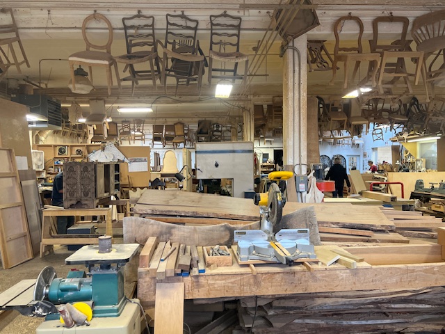 A workshop has unfinished chairs hanging from the ceiling and an assortment of woodworking tools.