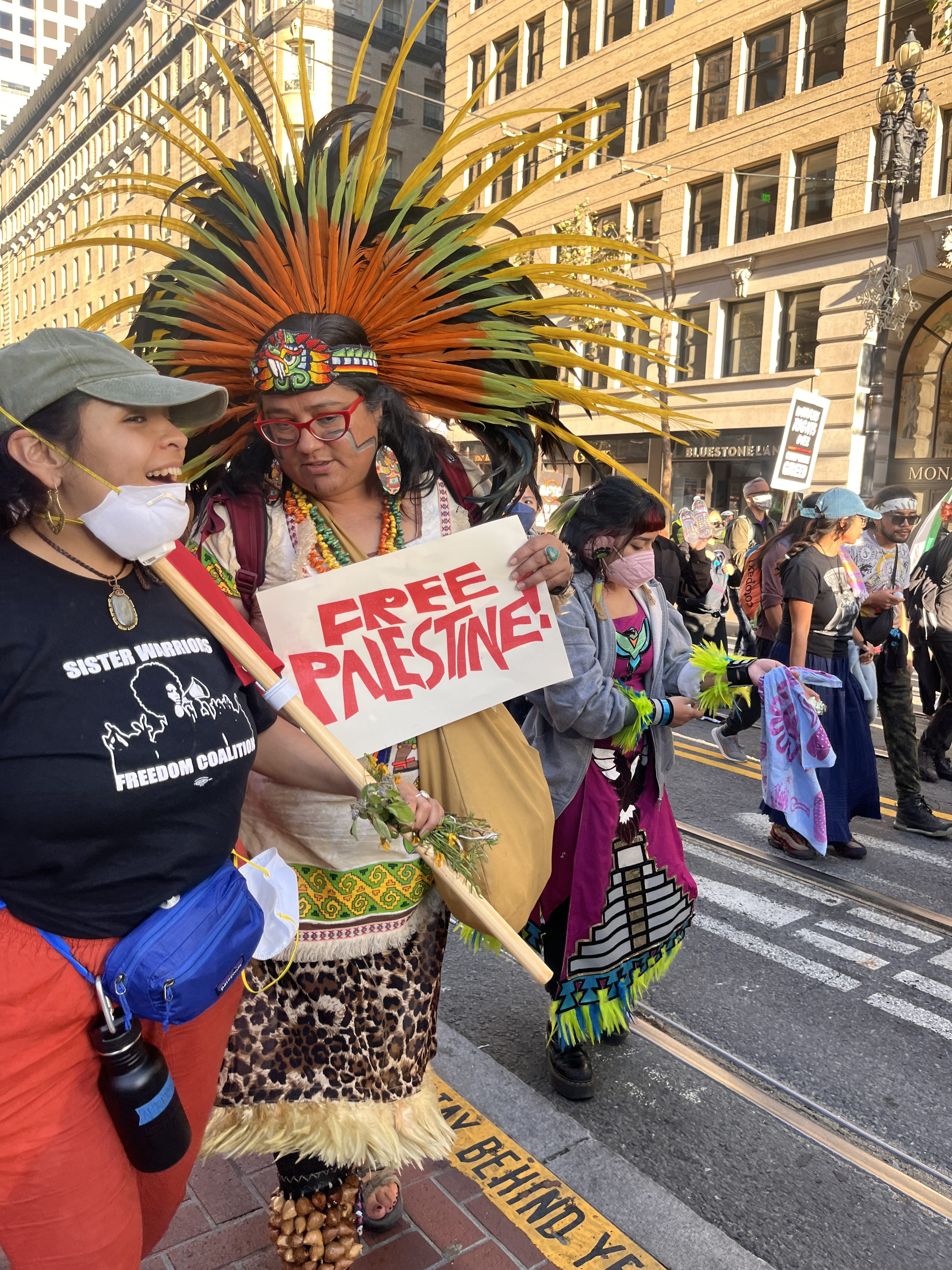 A woman in a feather headdress carries a sign reading &quot;Free Palestine&quot; while marching down the street.