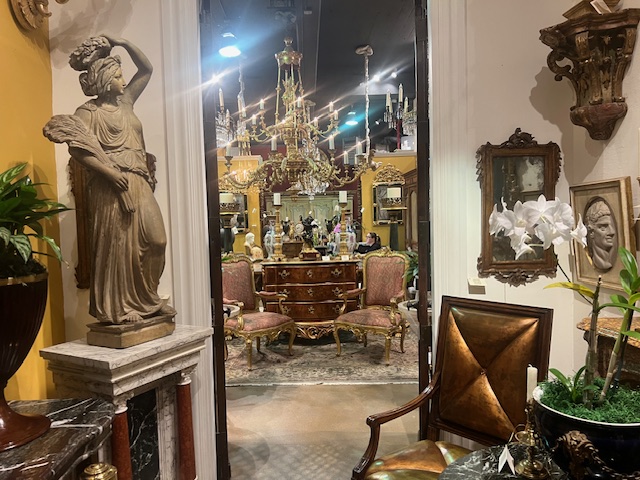 A showroom is stuffed with antiques, with a similarly decorated room visible through a door.