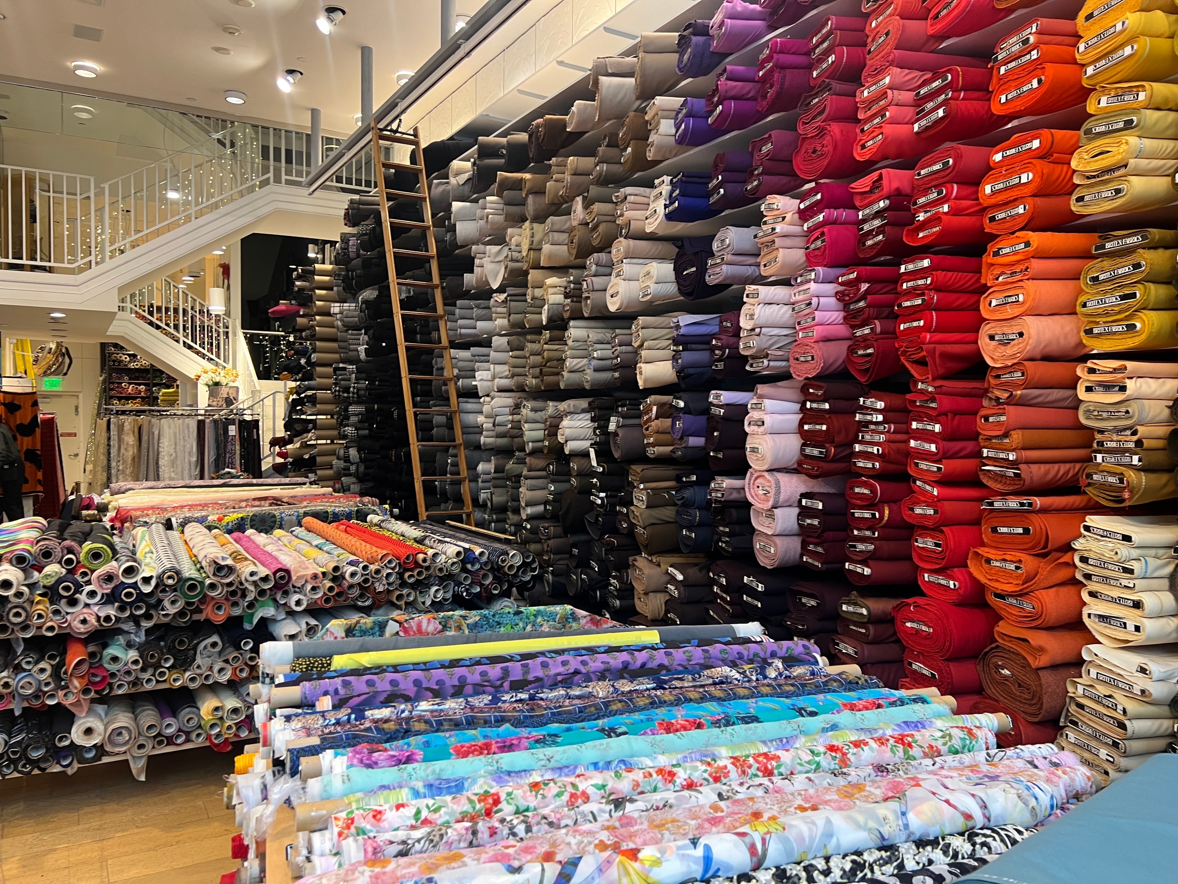 A wall is crowded with bolts of fabric that are organized by color, ranging from black and gray to purple, red and yellow.