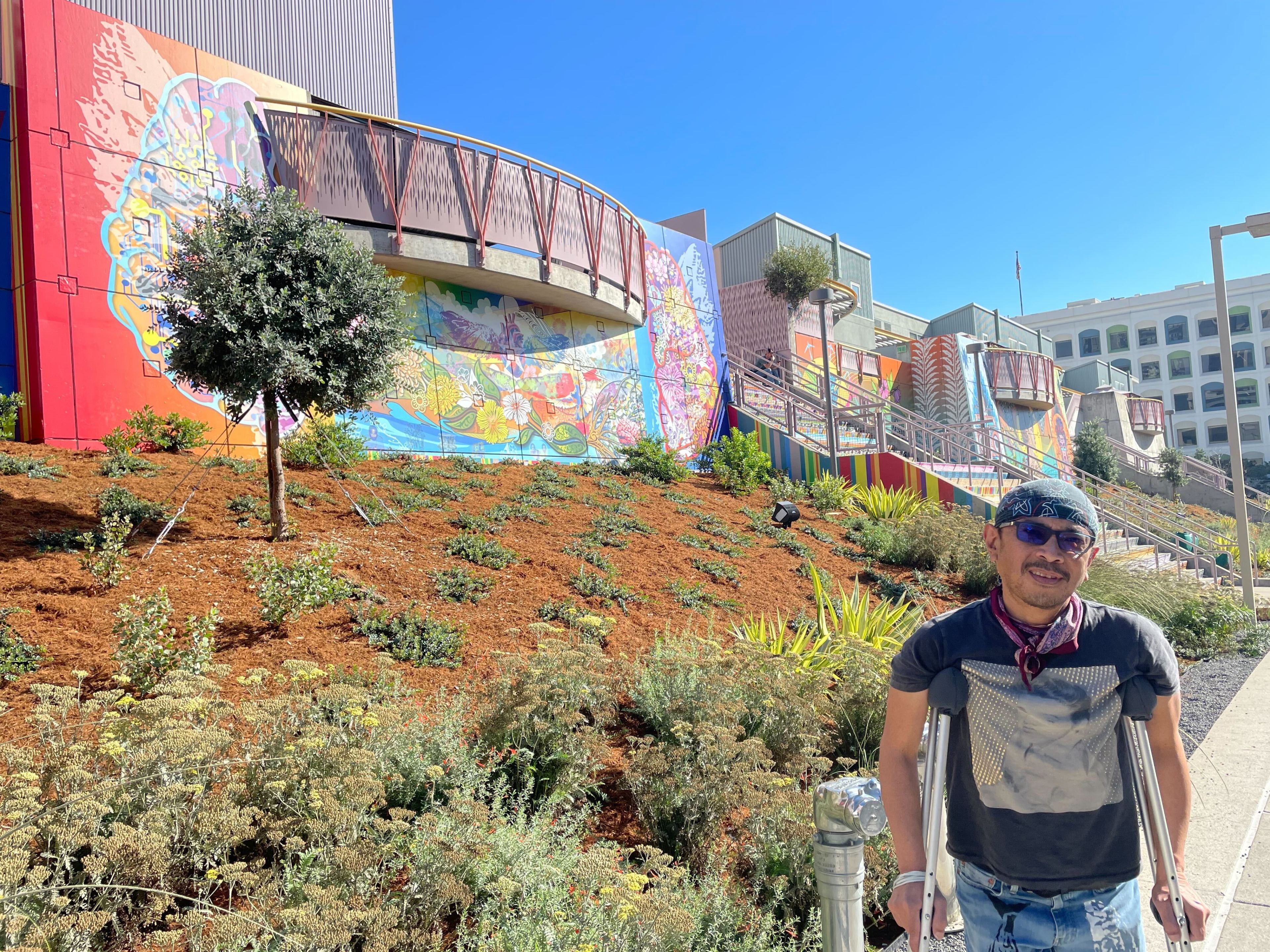 Artist Mel Vera Cruz, wearing a t-shirt, bandana and standing with crutches, stands in front of a bank covered in woodchips with low-lying scrubs planted sparsely throughout. Behind him is a mural Vera Cruz designed, depicting a stylized human brain.