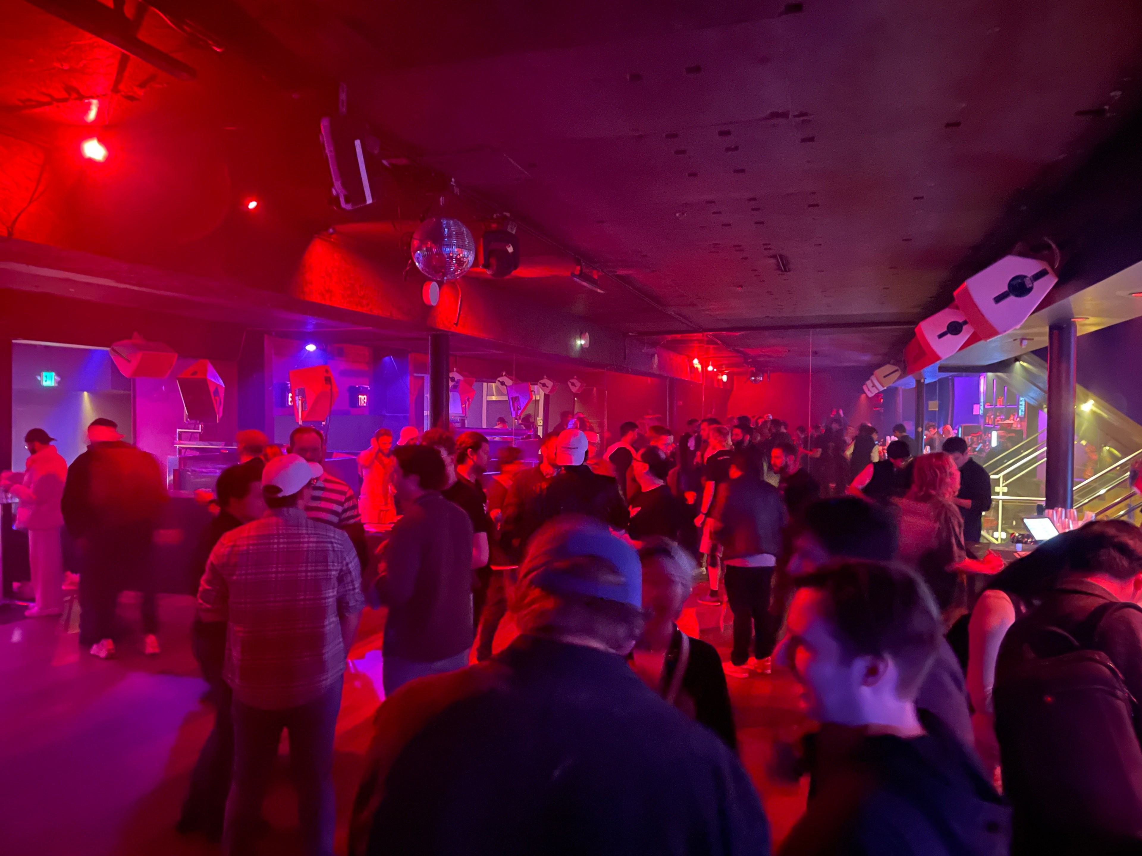 A large crowd of more than 100 people are seen standing with their backs turned and bathed in red and blue lights near a bar inside a nightclub with large speakers hanging overhead.