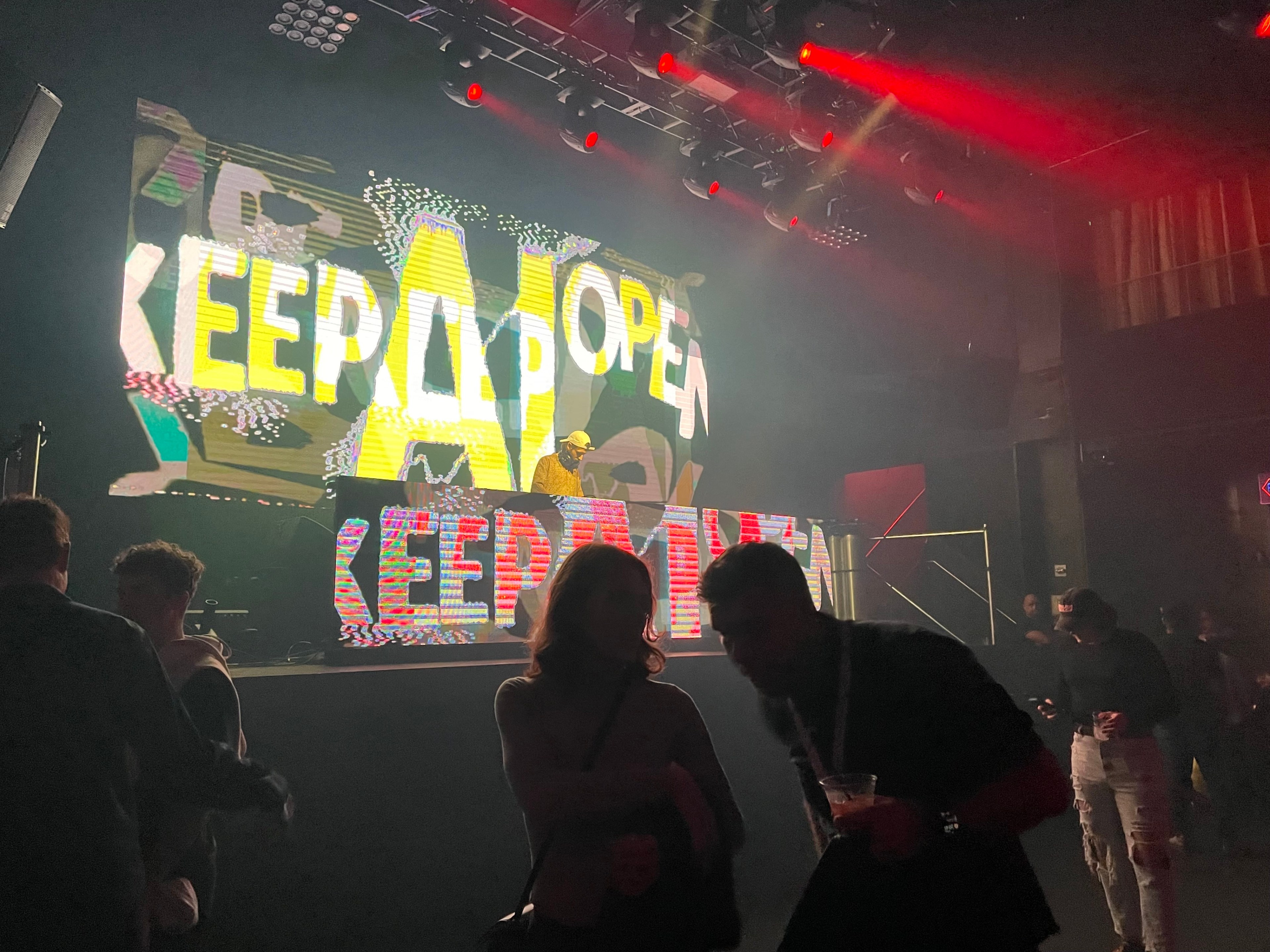 The silhouettes of several people are seen in front of a large light-up sign reading "keep AI open" in multi-color letters above a stage at a nightclub with spotlights shining red lights away from the stage.