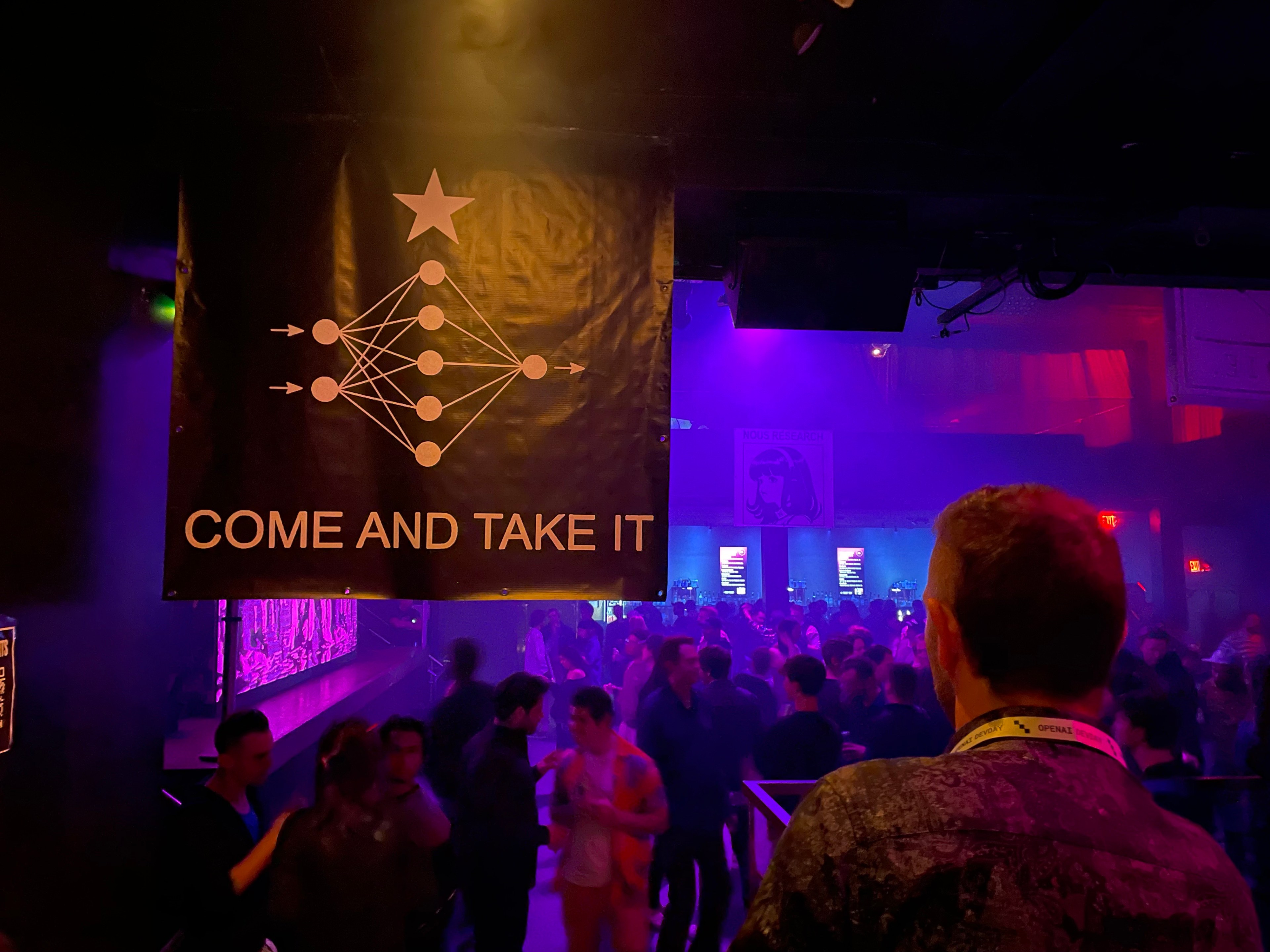 A person stands in the foreground to the right of a black poster reading "come and take it" next to an image with a series of dots connected with lines forming the web—a depiction of a "neural net" used by AI algorithms. Behind the poster is a crowd of people near a stage at a nightclub bathed in blue and purple light.