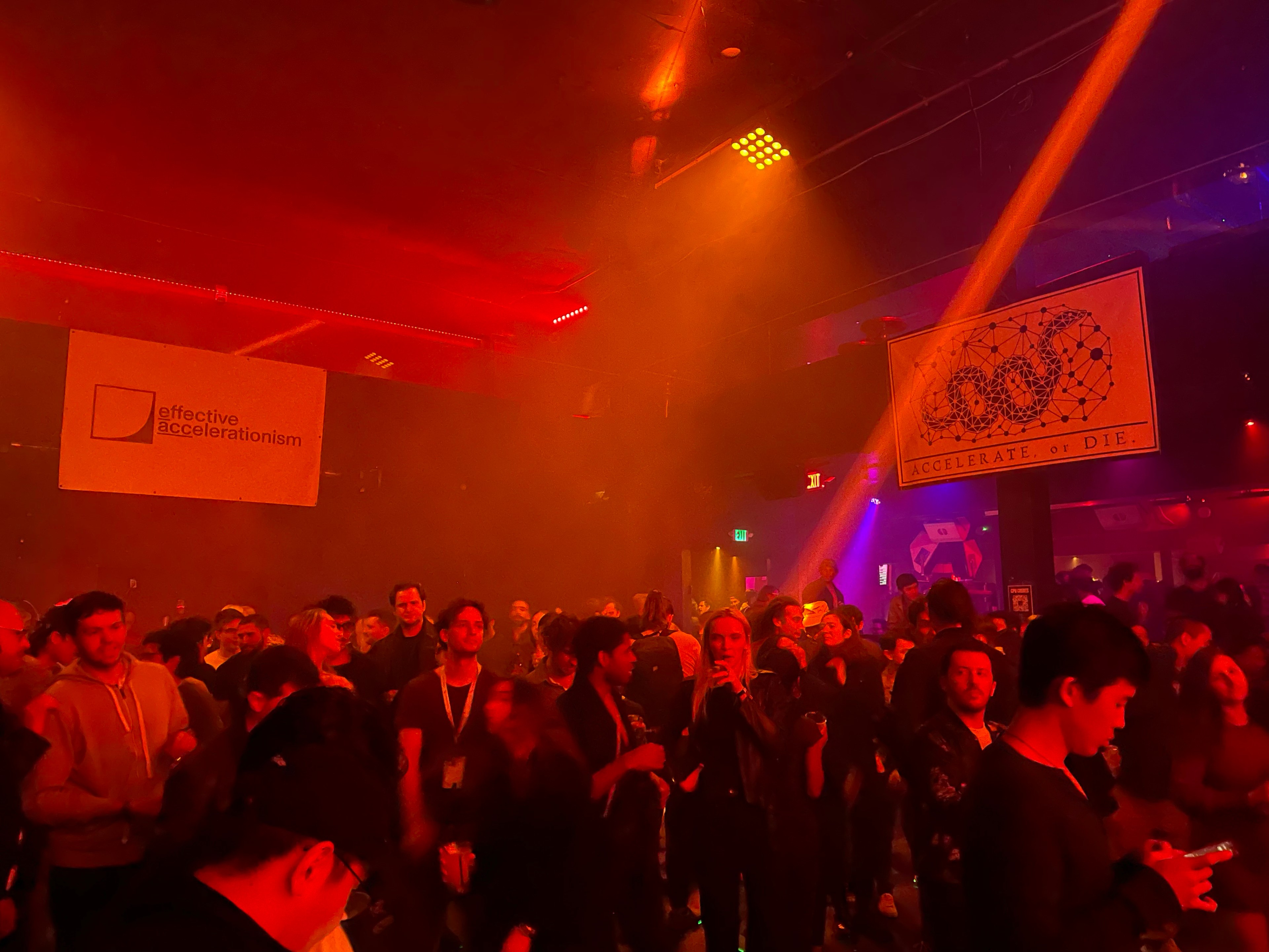 Hundreds of people stand on a nightclub dance floor bathed in red light with white posters reading &quot;effective accelerationism&quot; and &quot;accelerate or die&quot; hanging above them.