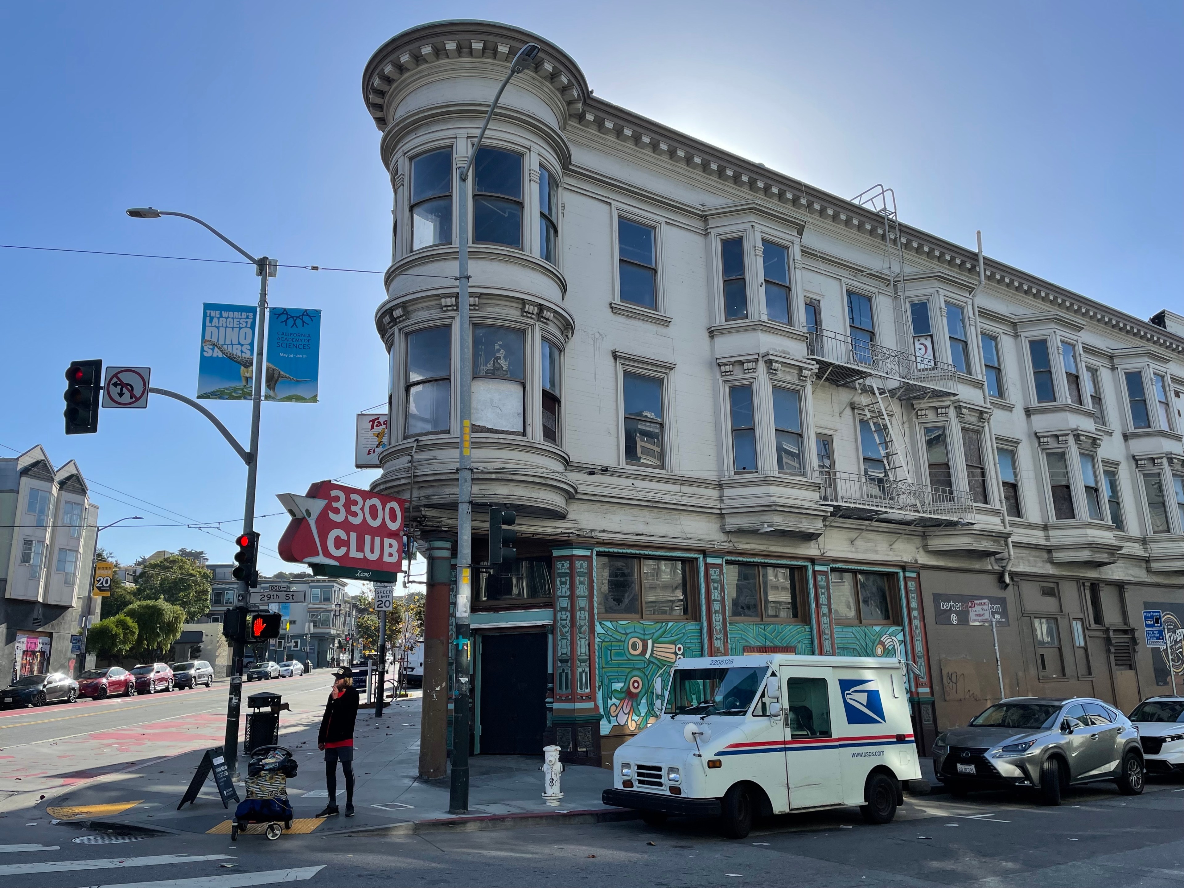 A vacant, gray three-story building sits on a San Francisco street corner, with a blade sign hanging over the sidewalk reading &quot;3300 Club,&quot; the name of the former bar there that burned down there in 2016. A man faces the viewer as he stands near the building waiting to cross the street, and on the right side of the photo a mail truck and sedan sit parked outside the vacant building.