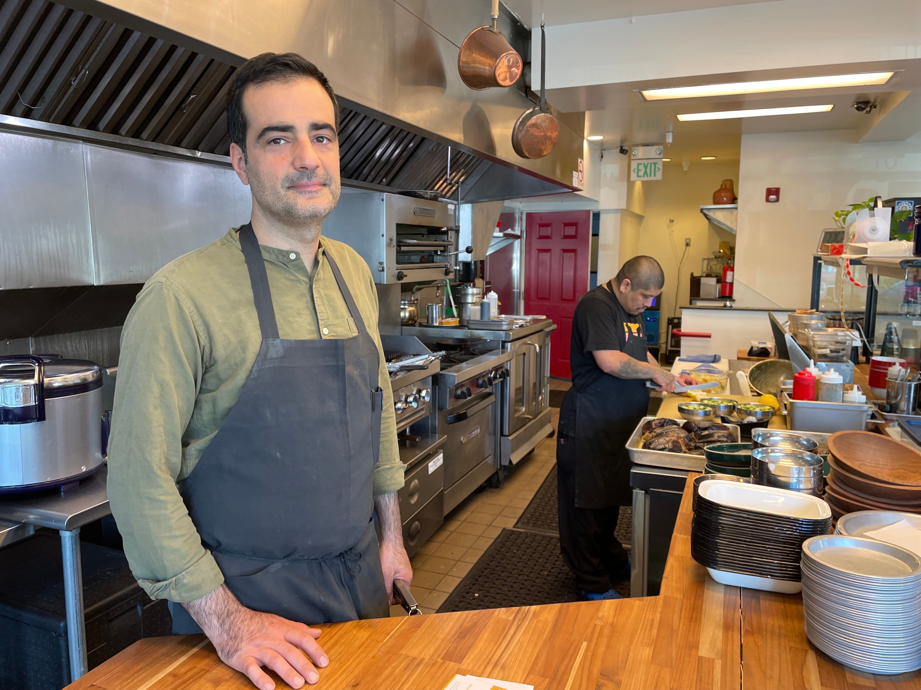 A man in a green shirt and grey apron stands with his hand on a wooden counter at a San Francisco restaurant with its kitchen and a sous chef chopping vegetables in the background.