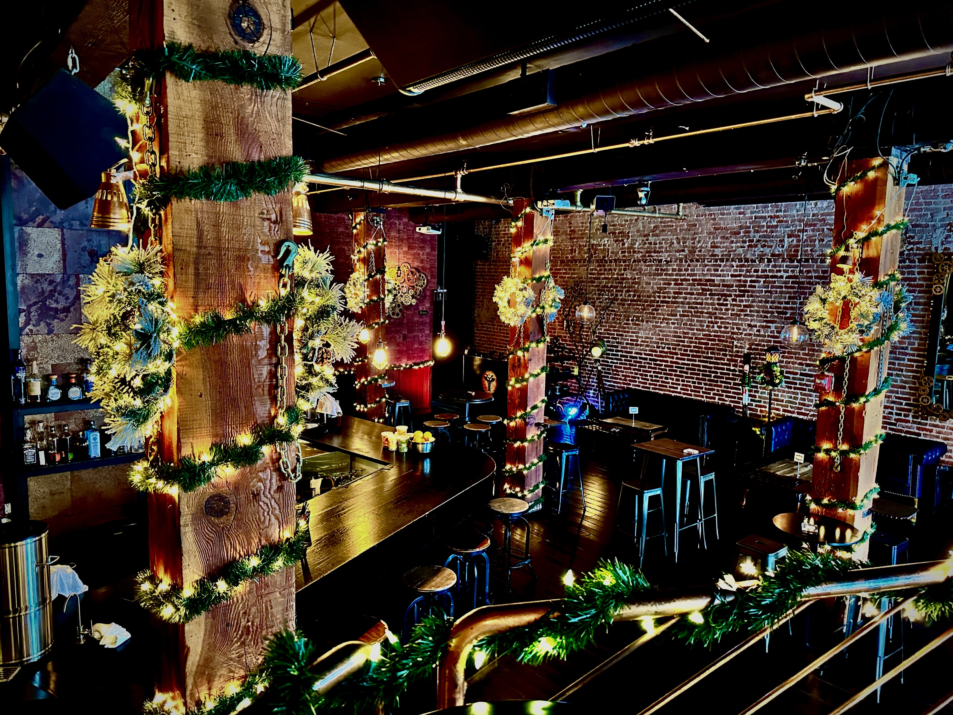Garlands and lights are wrapped around wooden pillars in an industrial-style bar with exposed brick walls. 