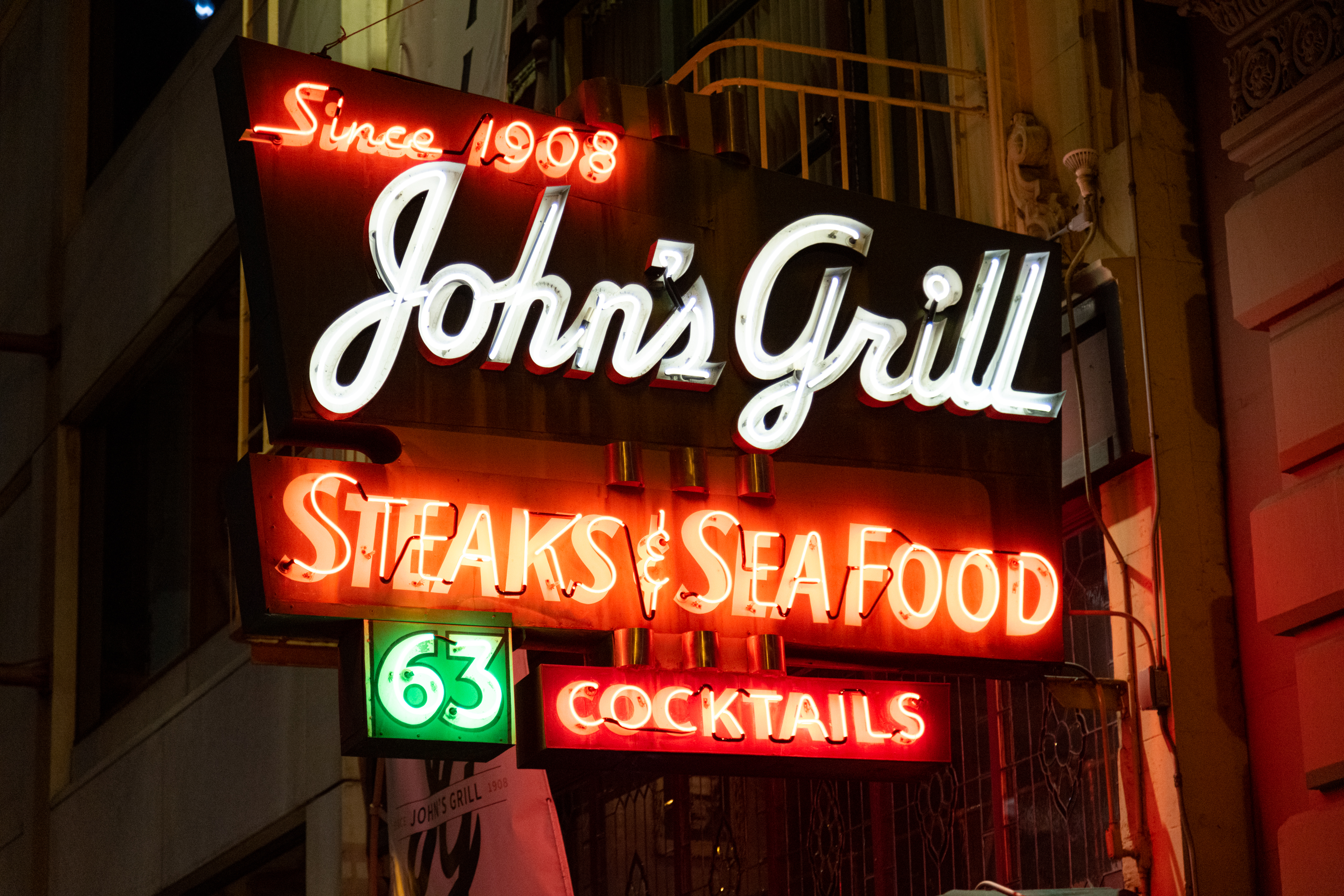 A multi-colored neon sign at night with the words &quot;Since 1908 John's Grill Steaks &amp; Seafood 63 COCKTAILS&quot;