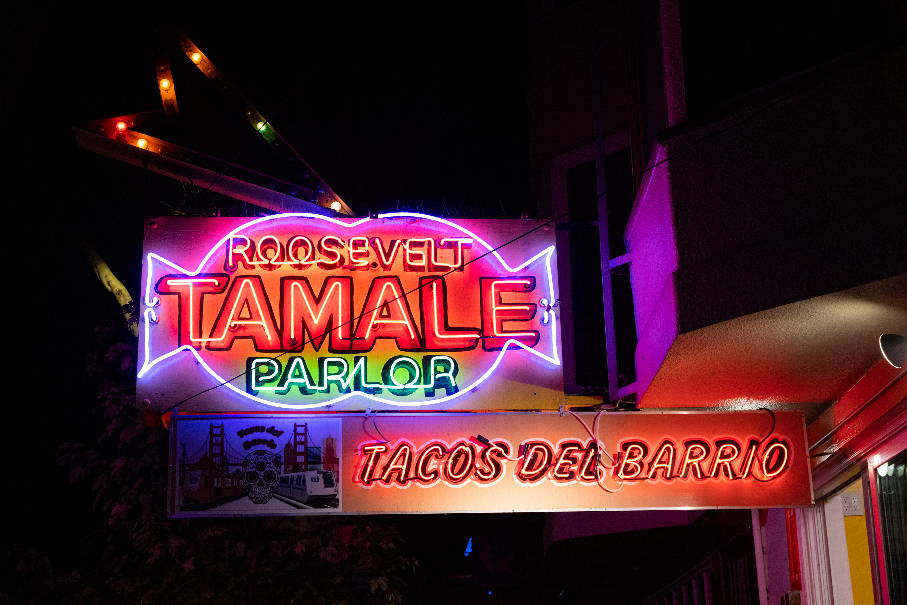A multi-colored neon sign at night with the words &quot;ROOSEVELT TAMALE PARLOR TACOS DEL BARRIO&quot;