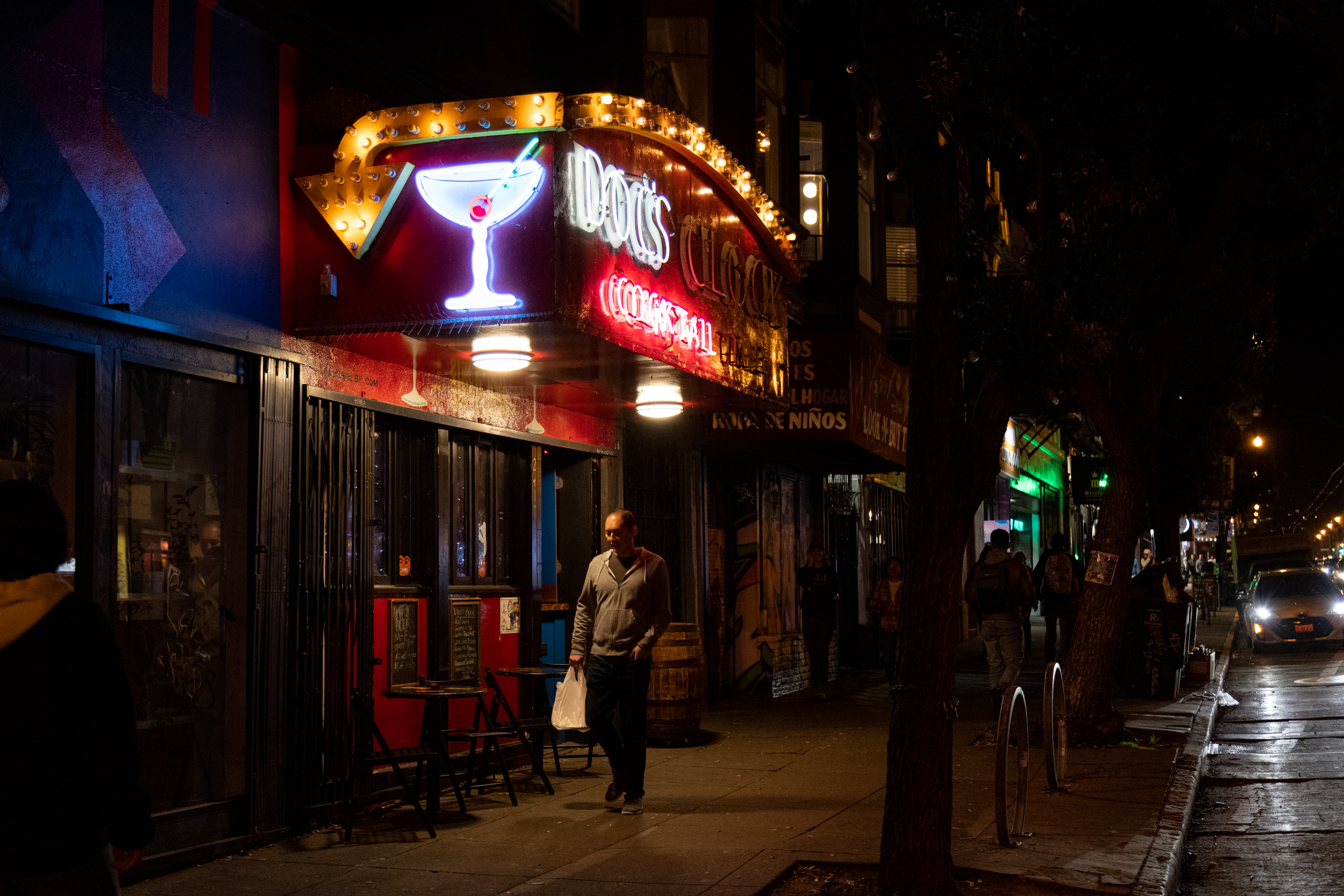 A street view of a person walking on the sidewalk below a neon sign that has a cocktail on it and reads &quot;DOC'S&quot;