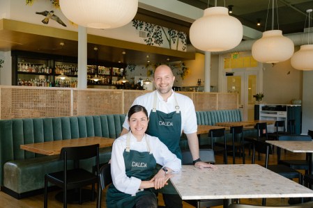 A smiling man and smiling woman, who are Executive chefs Laura and Sayat Ozyilmaz at Dalida, stand in the center of a modern restaurant that has large marble and wood tables spread throughout the dining space, circular lampshades on the ceiling and a bar in the background.