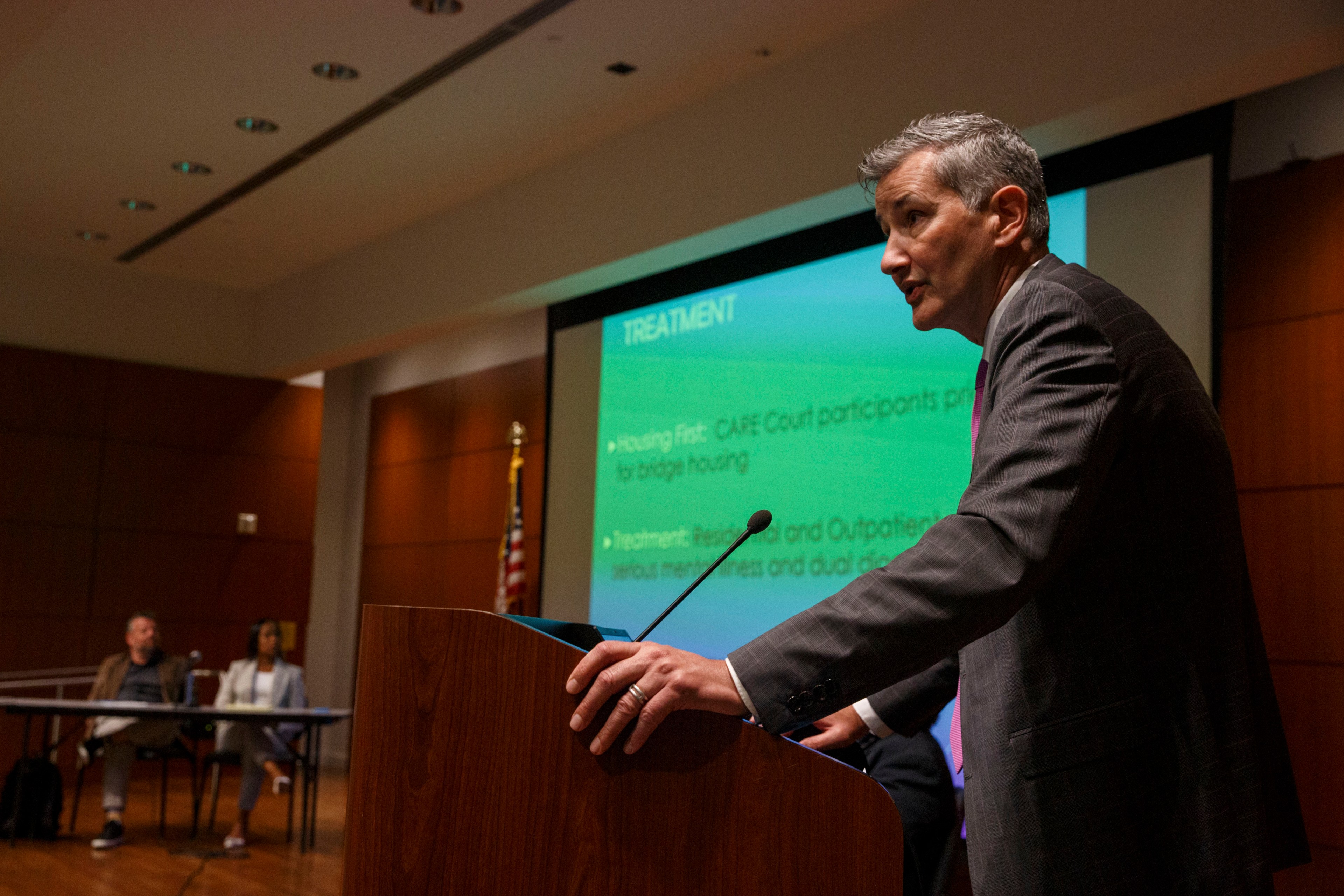 A man stands behind a podium in a lowly lit room and speak into a microphone. A screen is in the background and a title card read &quot;Treatment - Housing first: Care Court participants ...&quot;