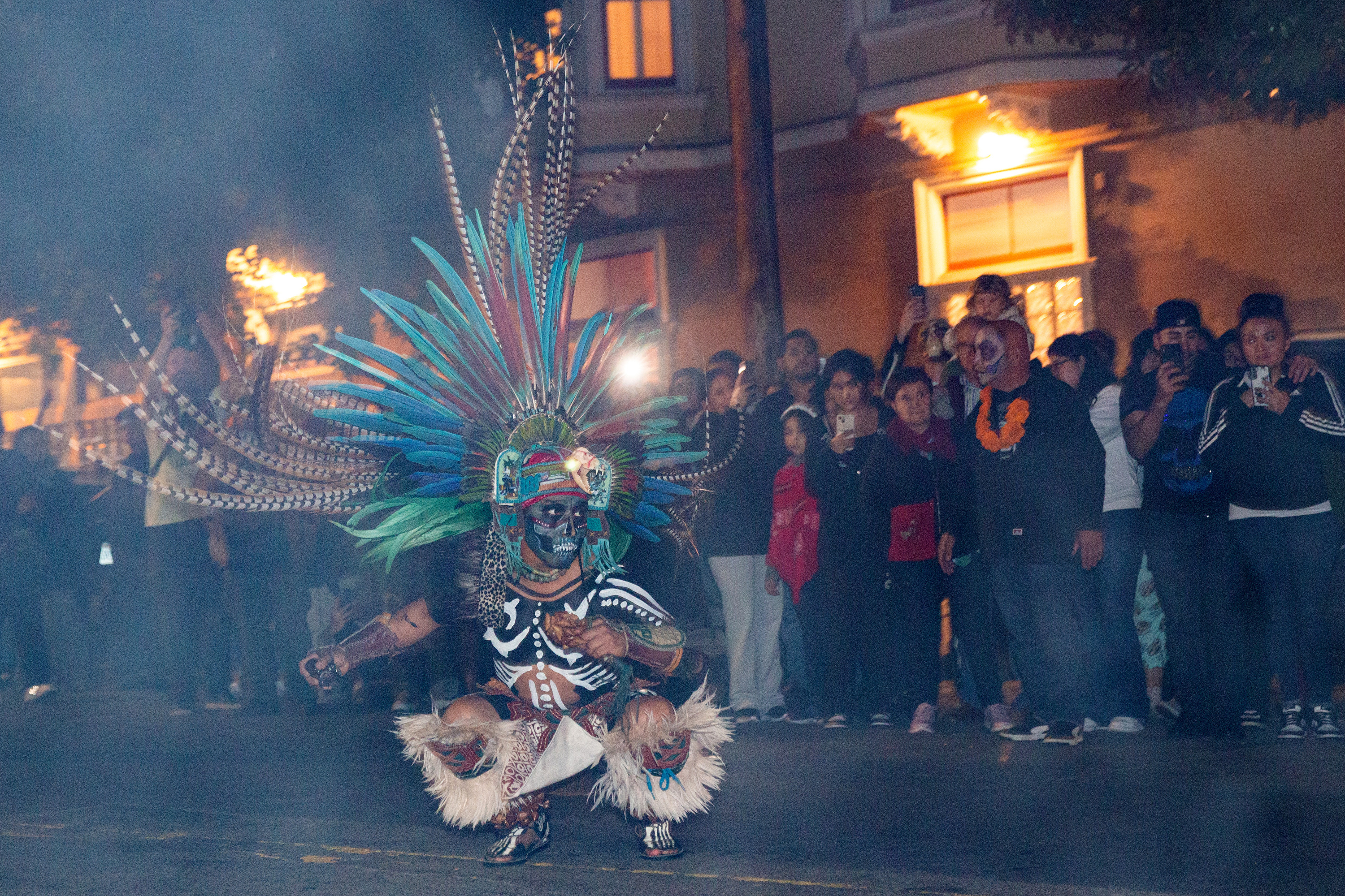 A person dressed in a feather headdress and costume performs on a smoky city street as people look on in the background
