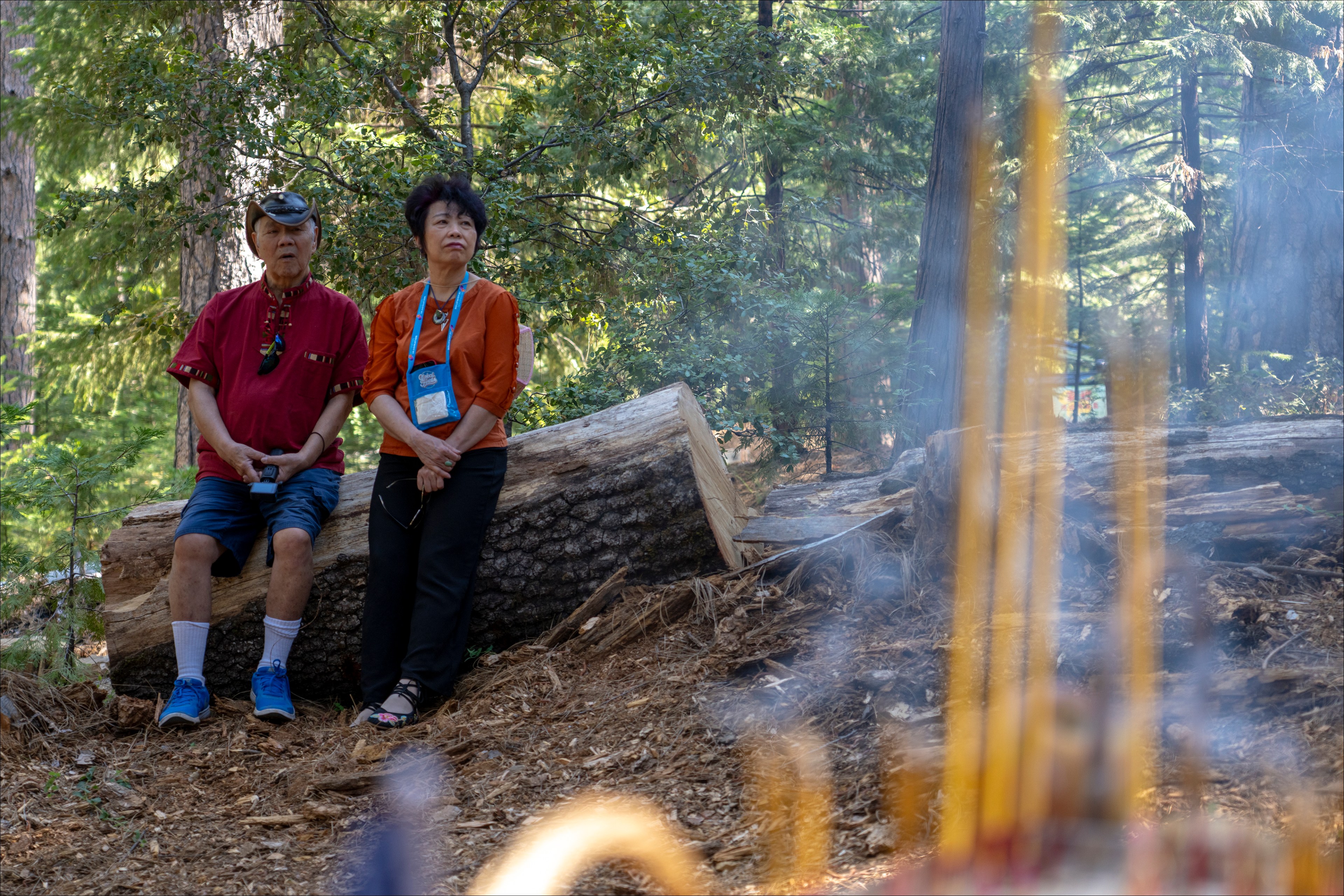 Two elderly people sit on a log in a forest with incense in the foreground.