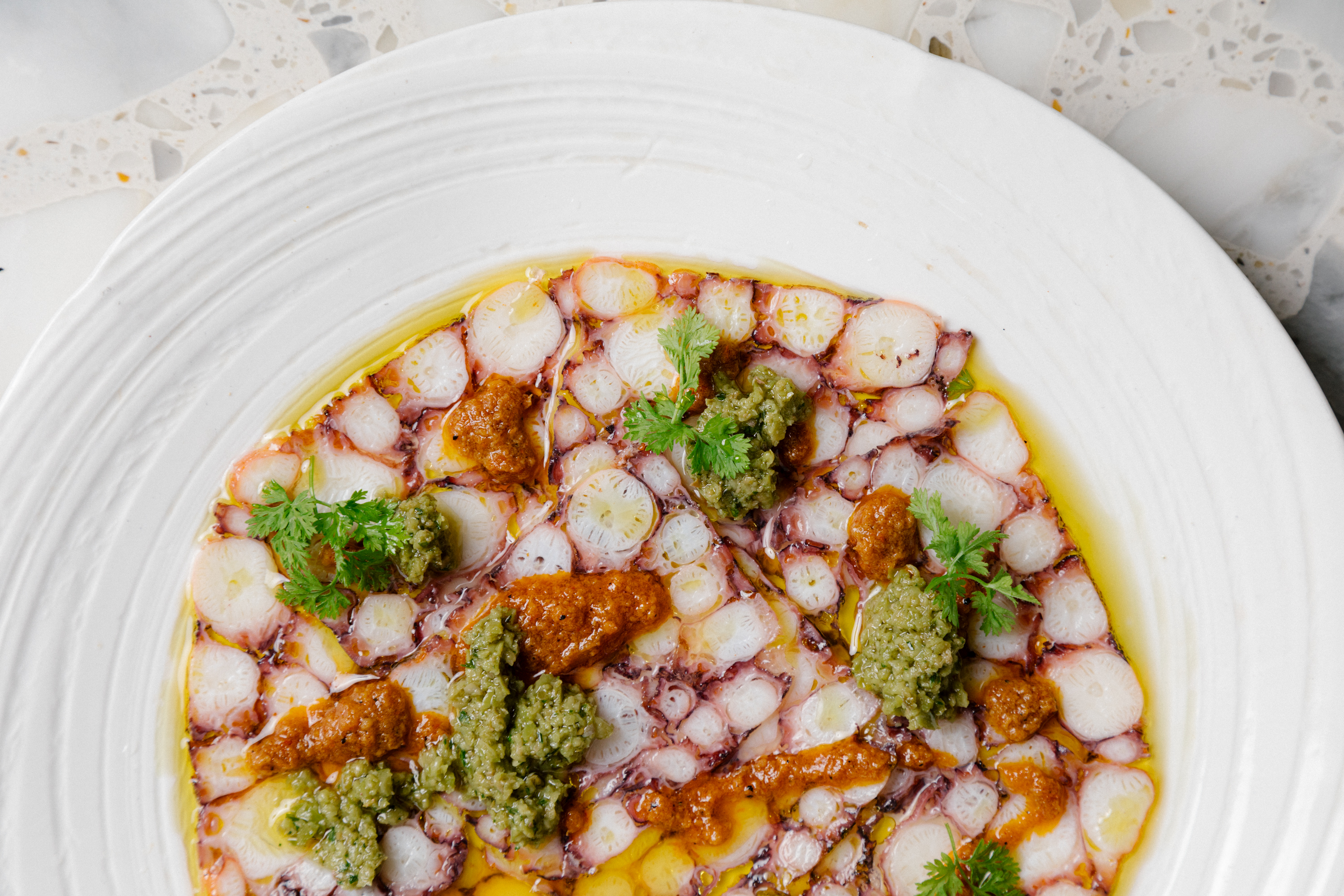 A close up photo of the dish &quot;Octopus and Sujuk&quot; which is filled with white and pink thinly sliced octopus, green olive-caper dressing and a yellow pork sujuk sauce in a ceramic white dish.