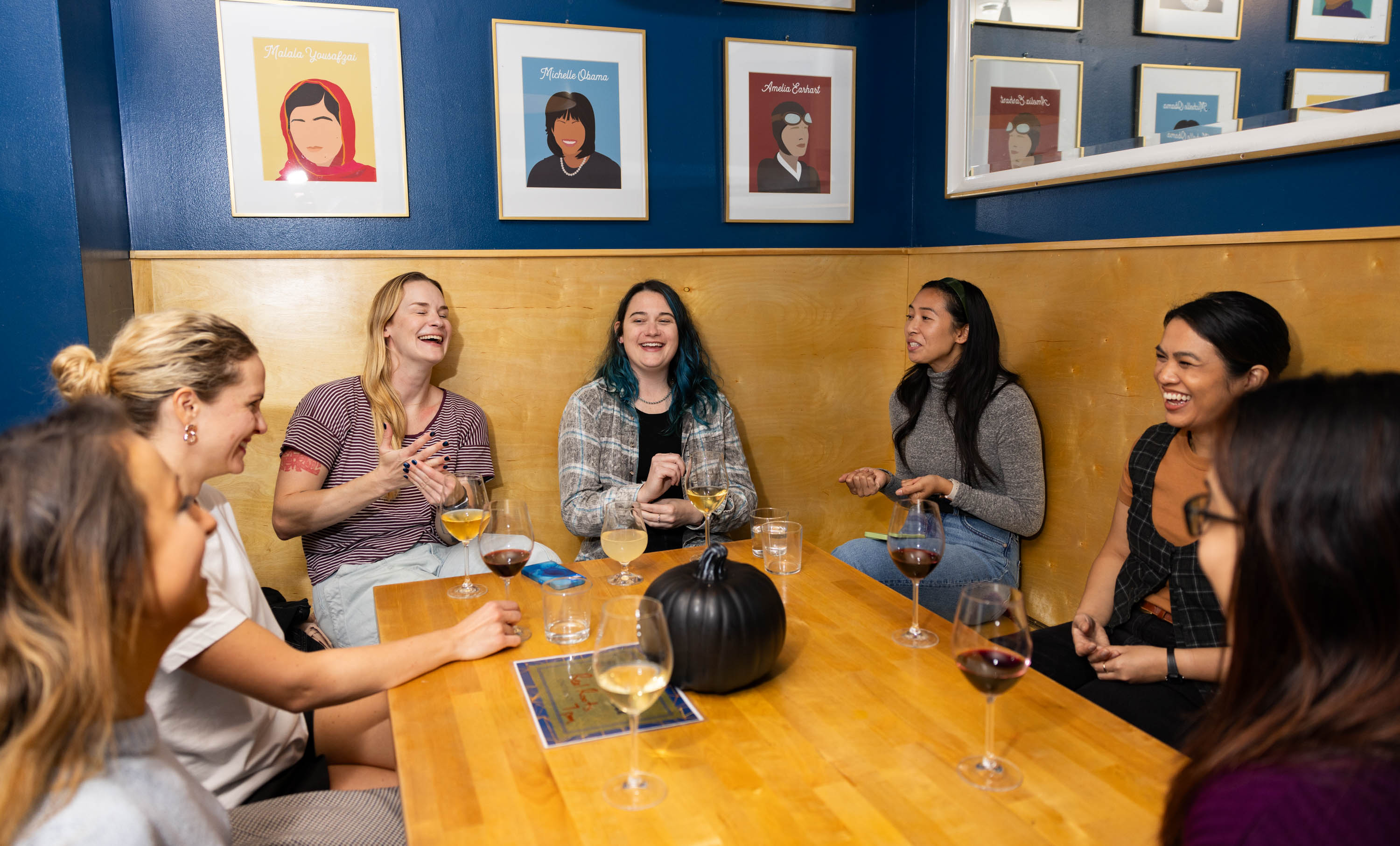 A group of women sit around a table smiling and laughing as wine glasses sit on the table in front of them.