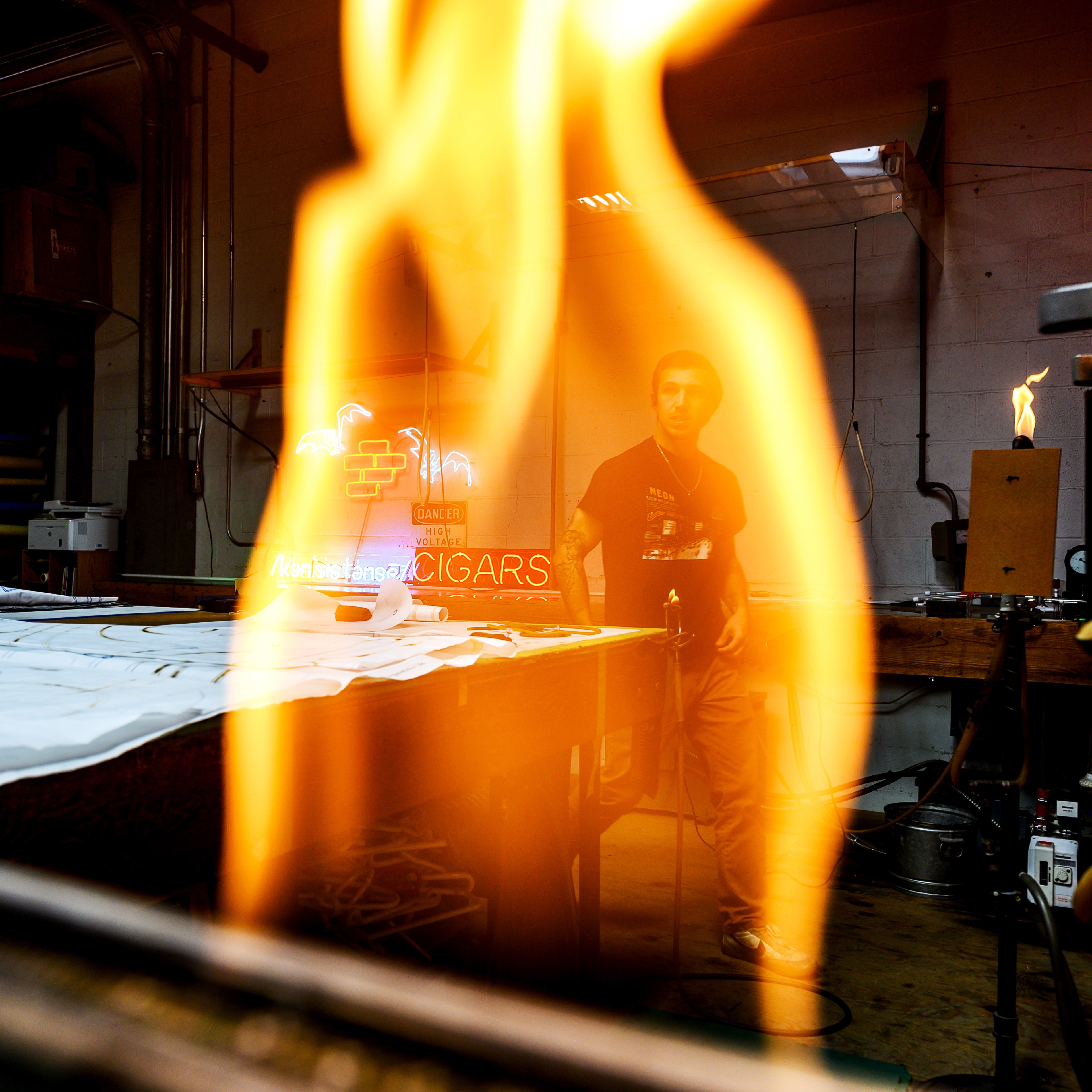 A large flame of fire in the foreground with a man in the background of a workshop.