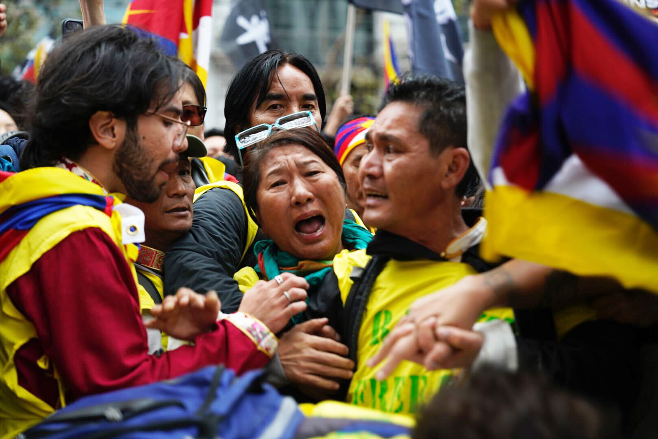 A woman screams while in the middle of dozens of people during a protest with Tibetan flags in the background.