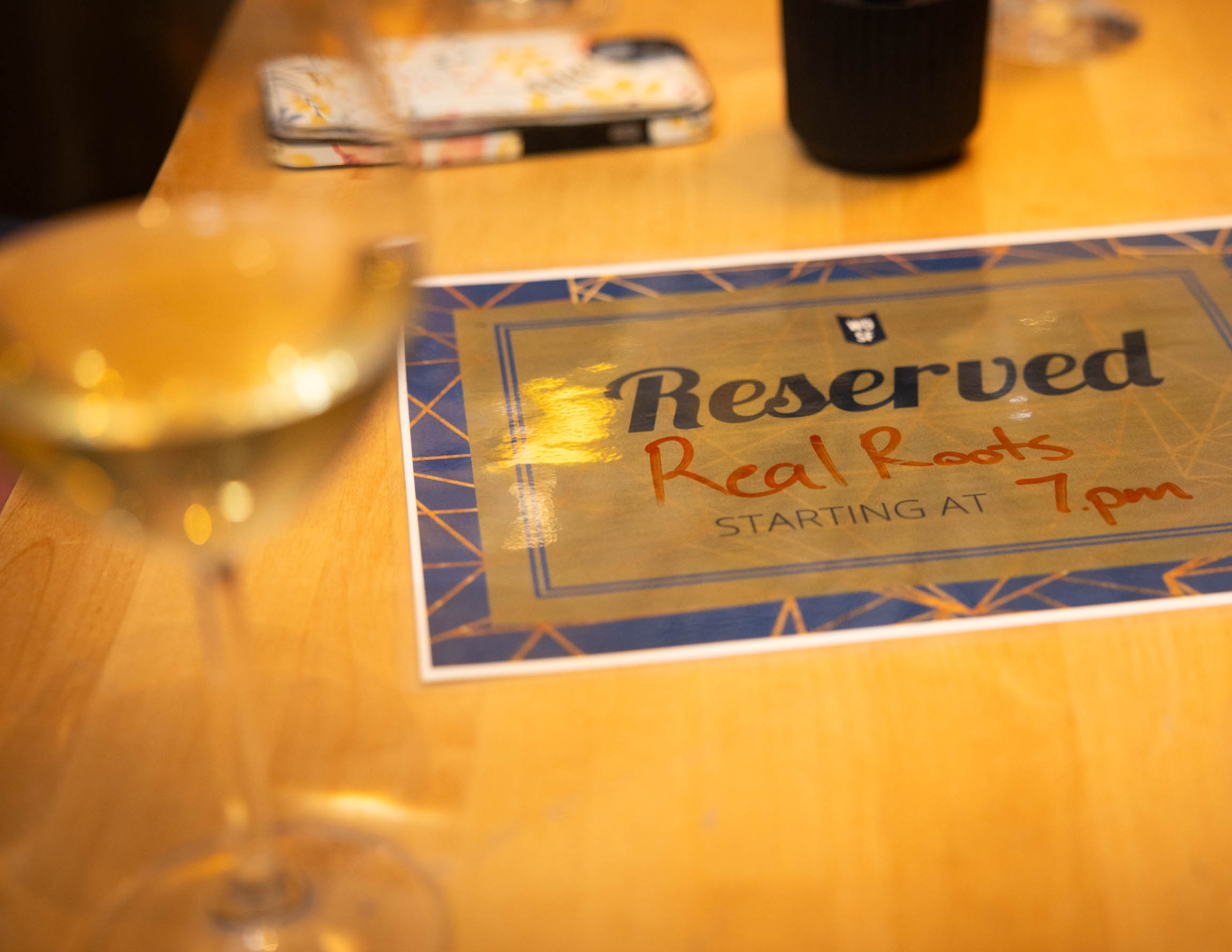 A tight photo of a piece of paper that reads &quot;Reserved Real Roots Starting at 7pm&quot; with an out of focus glass of wine in the foreground.