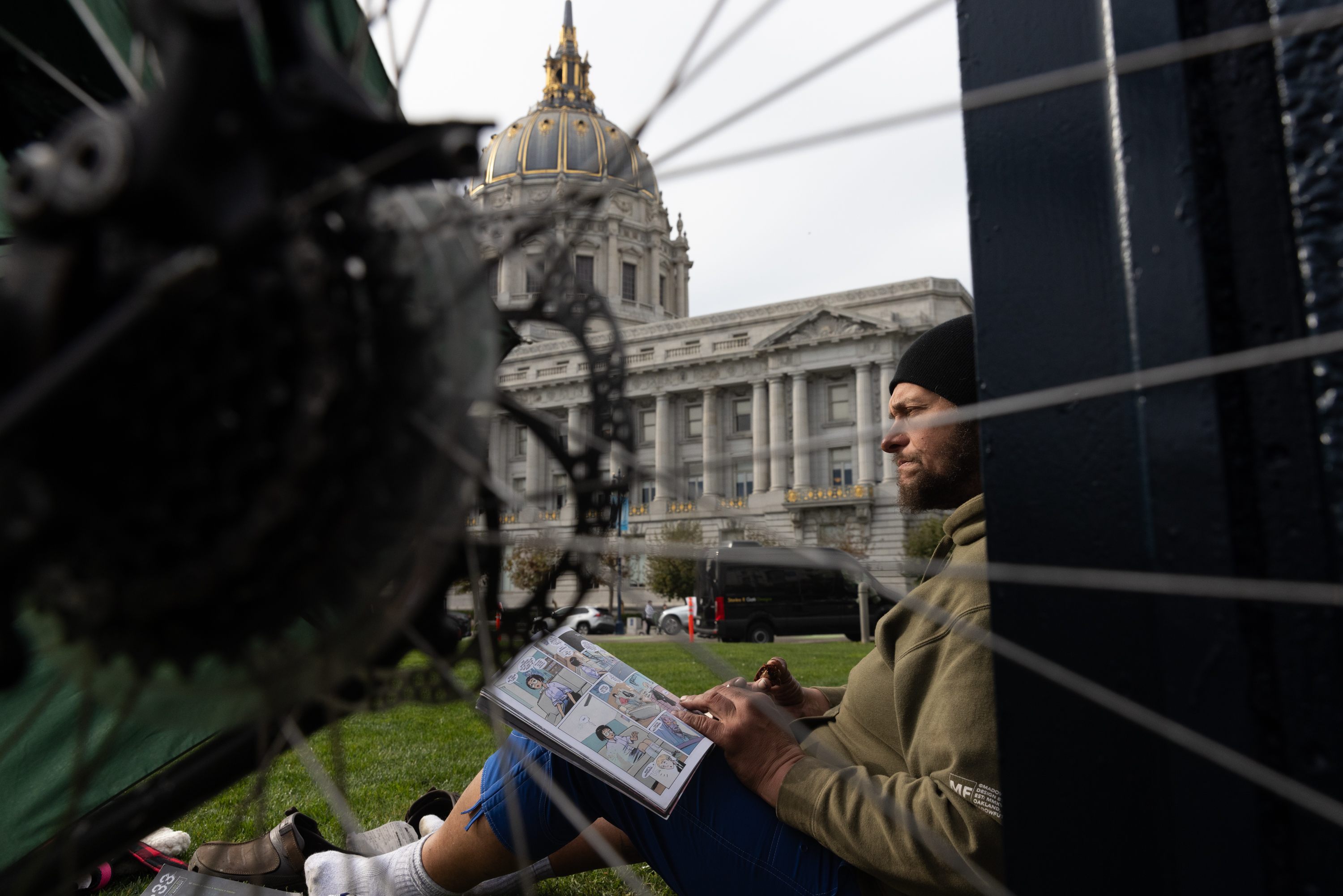 A man sits on the grass infront of City Hall and a bike wheel in the foreground.