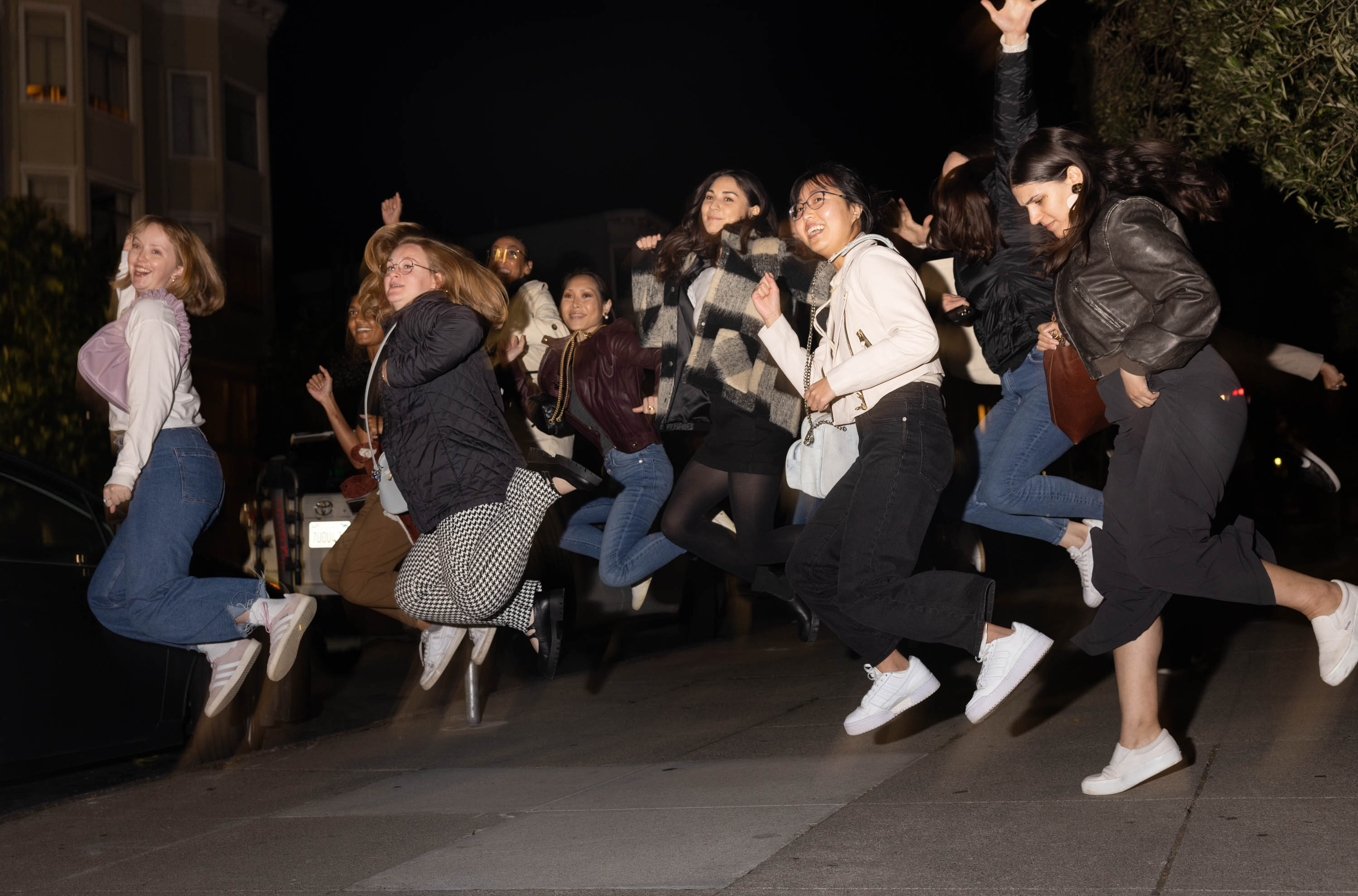 About one dozen young women jump in the air simultaneously while standing on the sidewalk at night.