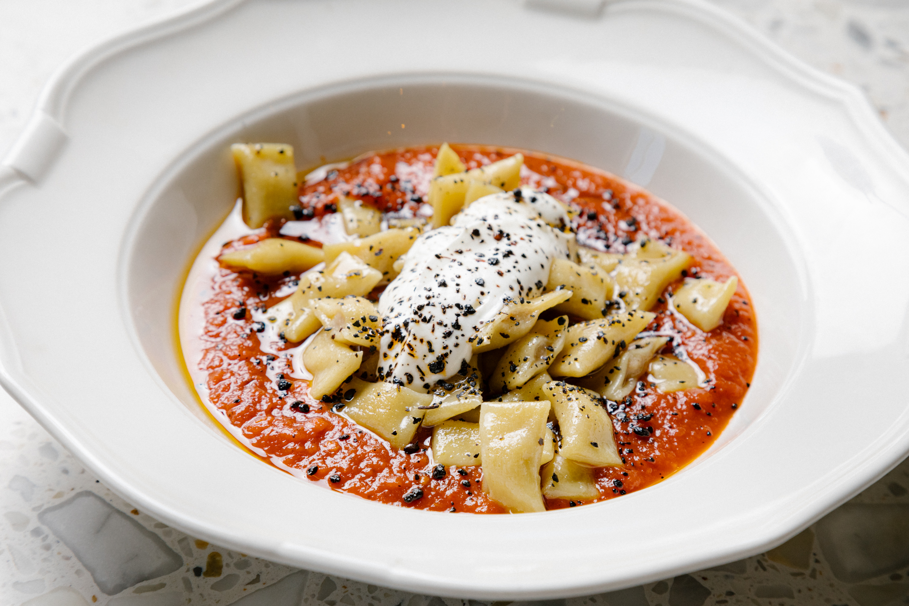 A close up photo of a pasta dish called “Kayseri Manti” which included small butter-roasted lamb pasta dumplings spread out on a white, ceramic dish with a large garlic yogurt in the center resting in red tomato sauce.