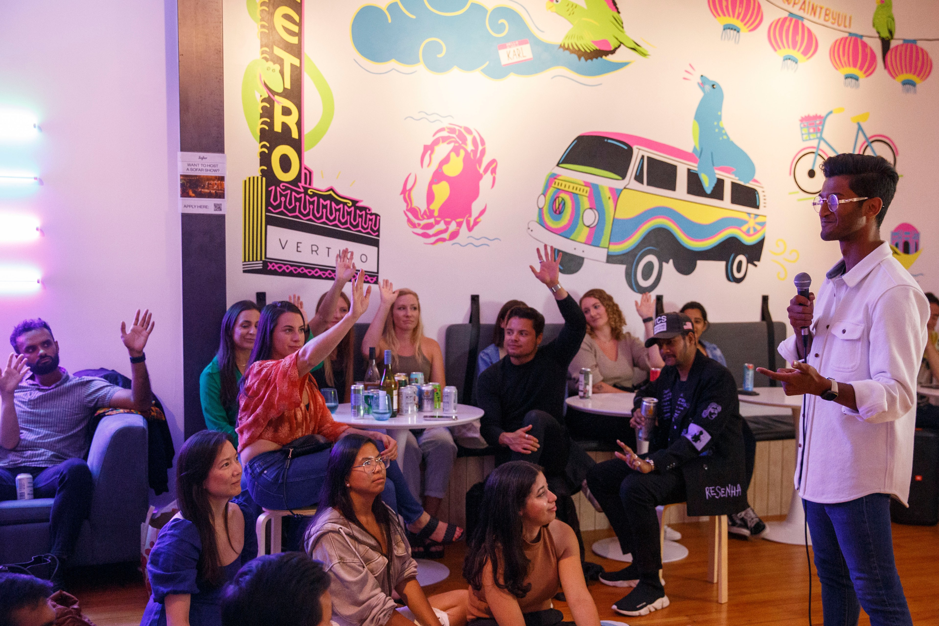 In a lounge space with fun wallpaper that includes a pink crab, a blue sea lion on a Volkswagon bus about a dozen people listen to a man holding a microphone who stands off to the right. Some of the people are seated on the floor and some are seated on chairs and roughly half the people are raises their hands in the air.