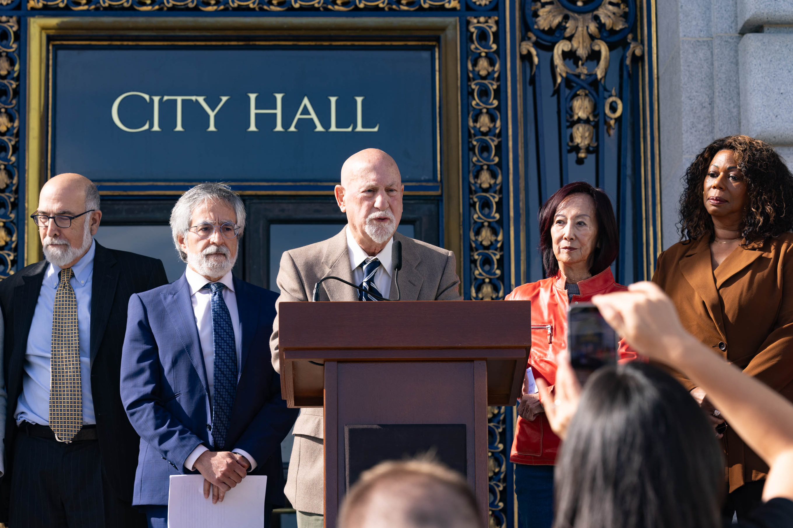 A man standing behind a podium speaks to an audience whit standing in front of a facade that reads &quot;CITY HALL&quot; as 4 other people stand beside him looking on.