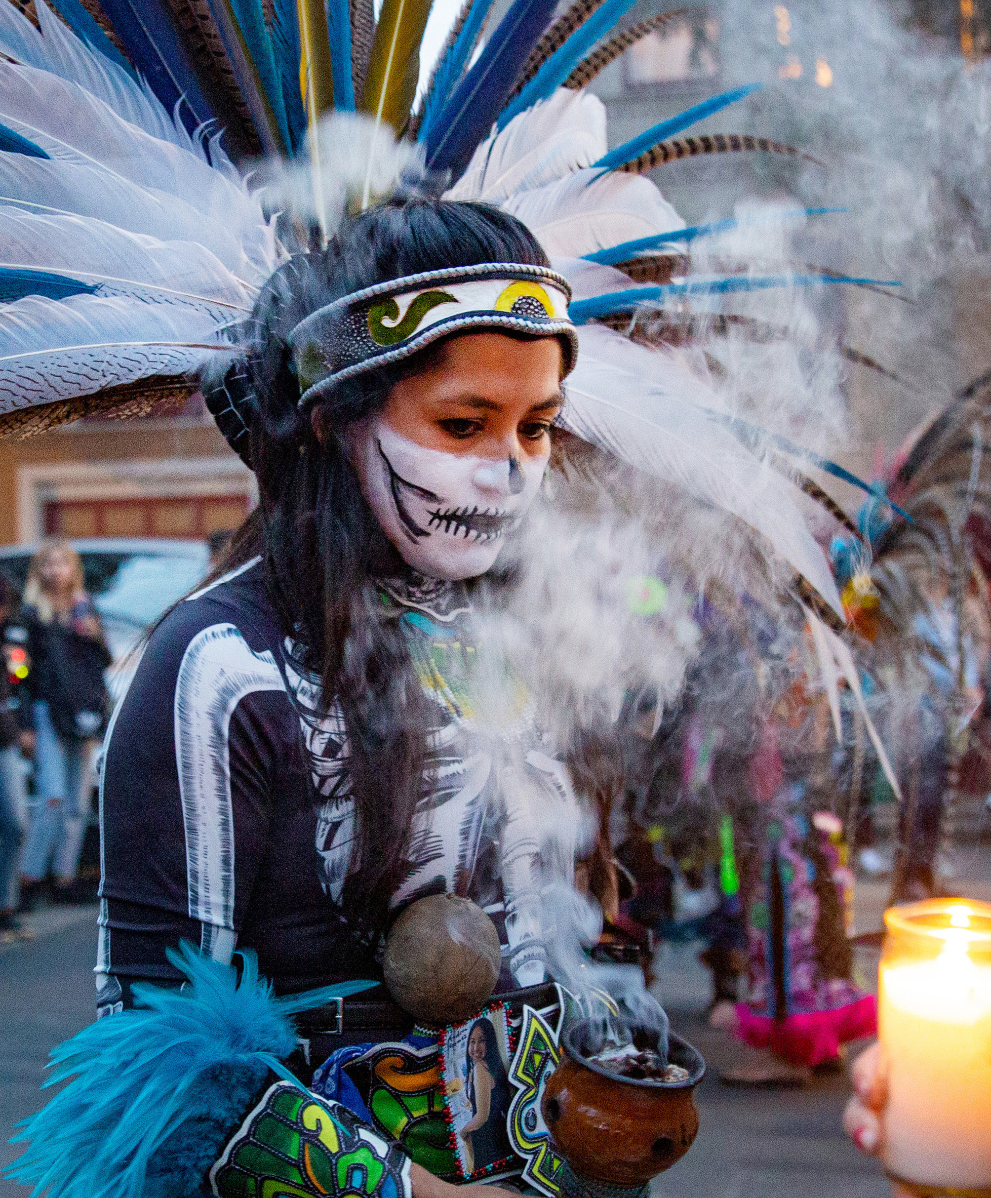 A person dressed in a feather headdress and costume burns incense during an outdoor event