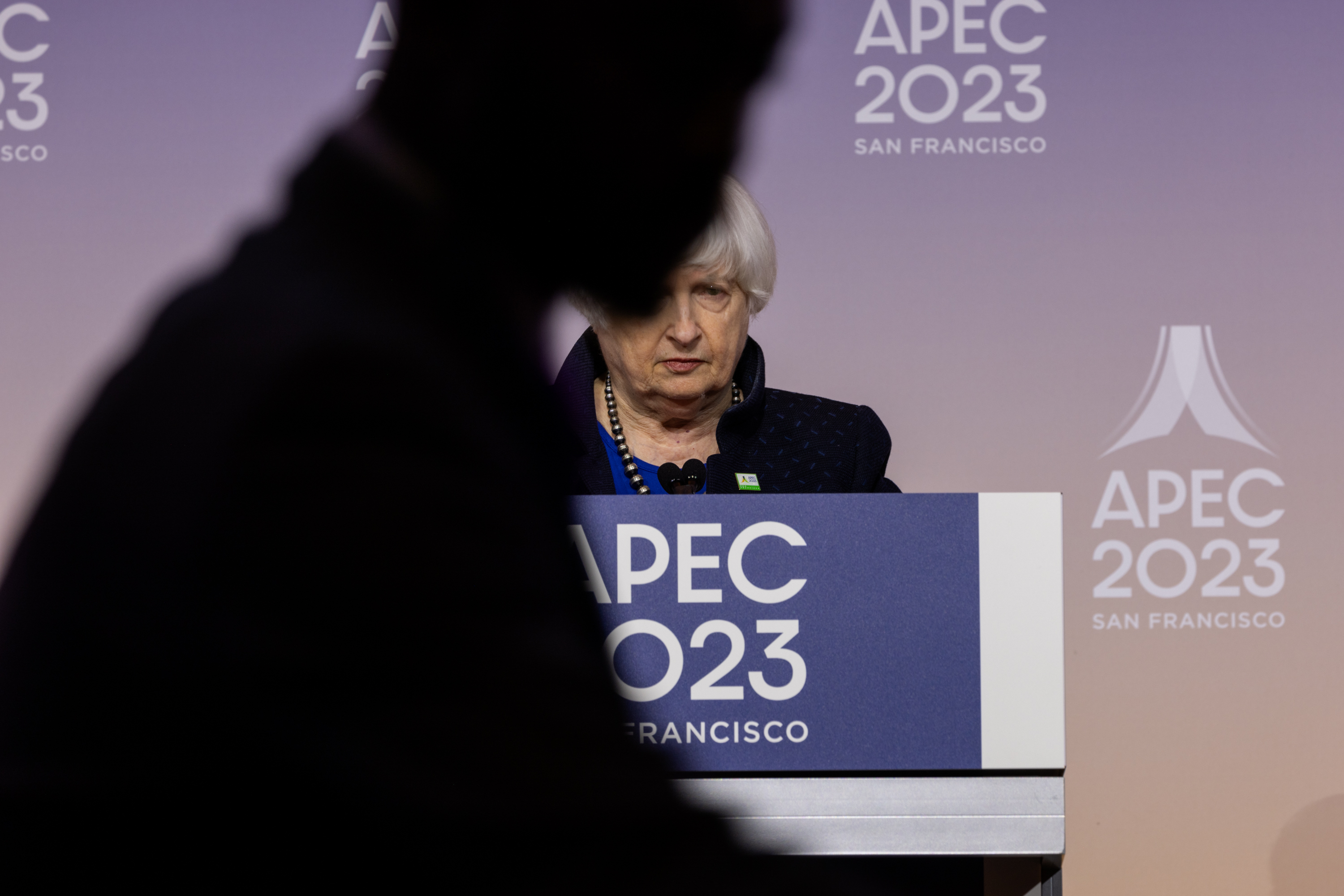 Janet Yellen, United States Secretary of the Treasury, stands behin a podium that read APEC 2023 as a silhouette of a person blocks a portion of her face.