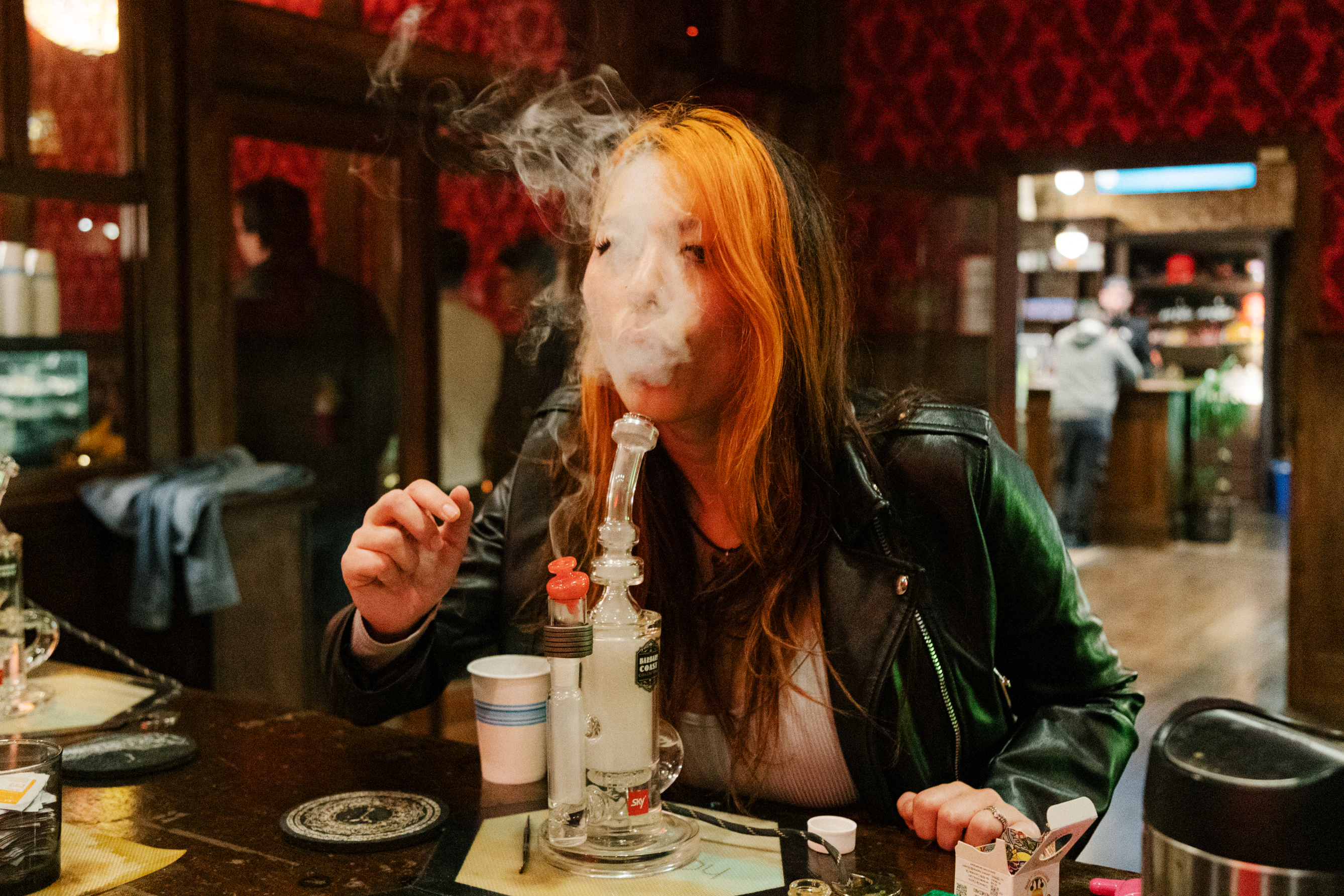 A woman blows out smoke after smoking out of a cannabis pipe in a cannabis dipsensary - some people are in the background shopping for cannabis merchandise.