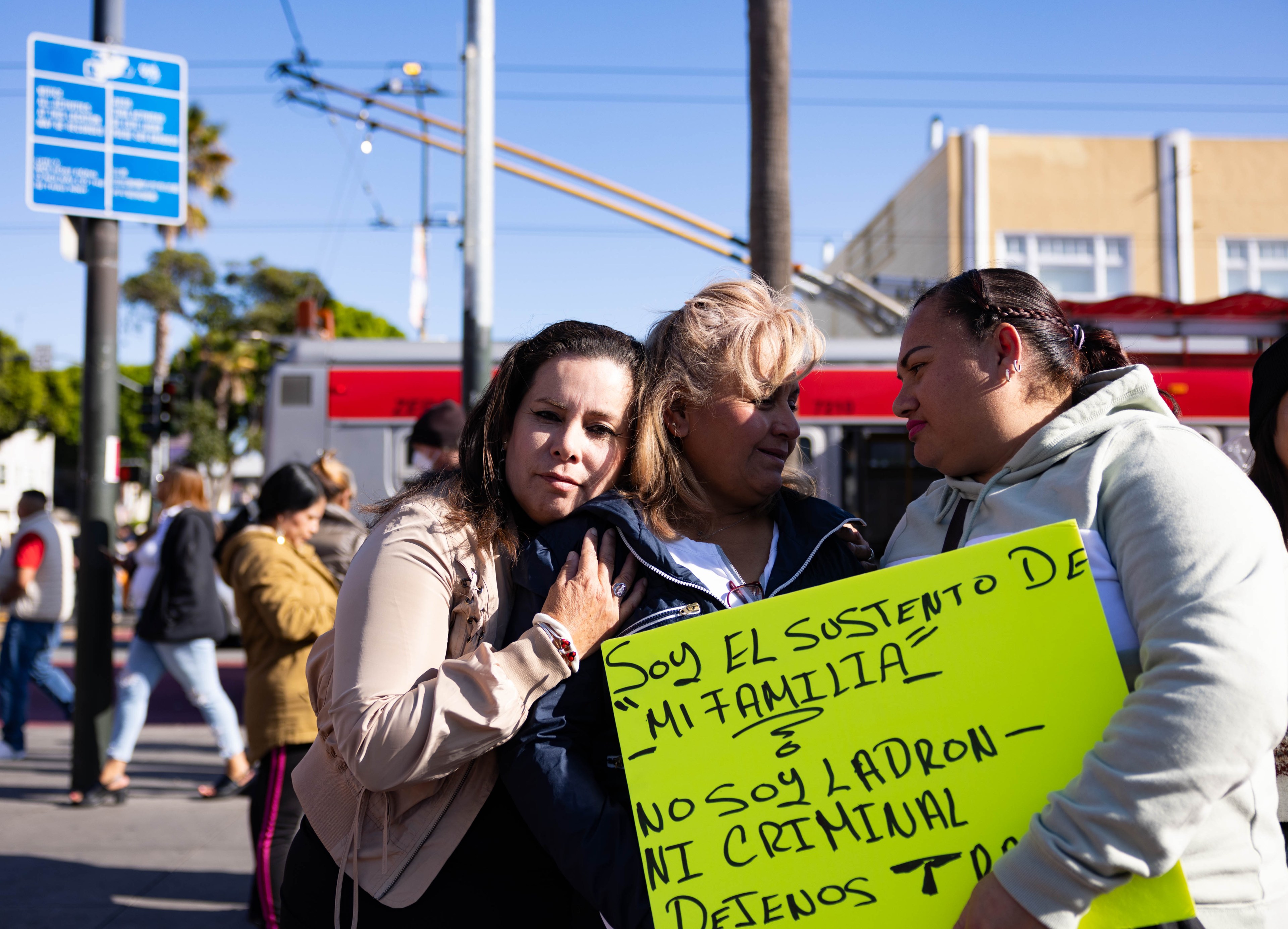 3 women comforting one another with a sign that reads "Soy el sustento De 'Mi Familia' - No Soy Ladon Ni Criminal ..."