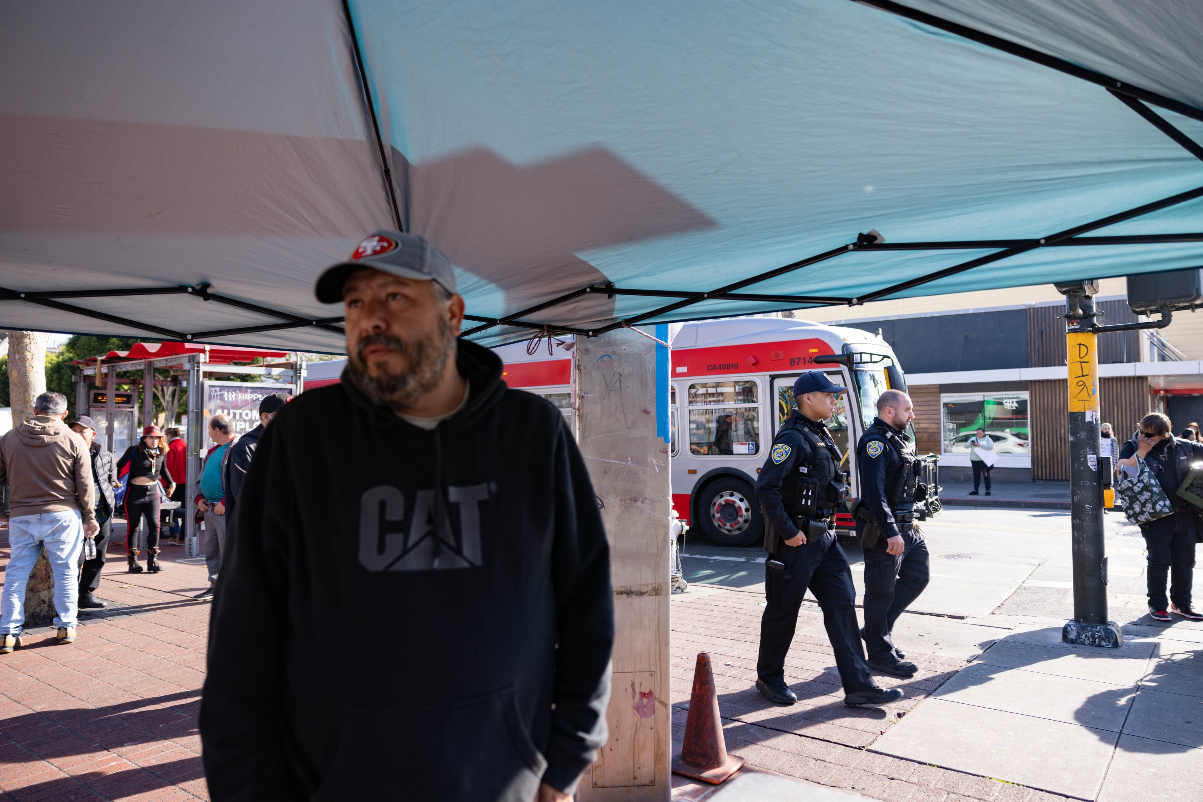 An out of focus man looks into the distance while standing under a tent with SFPD officers passing down the sidewalk.