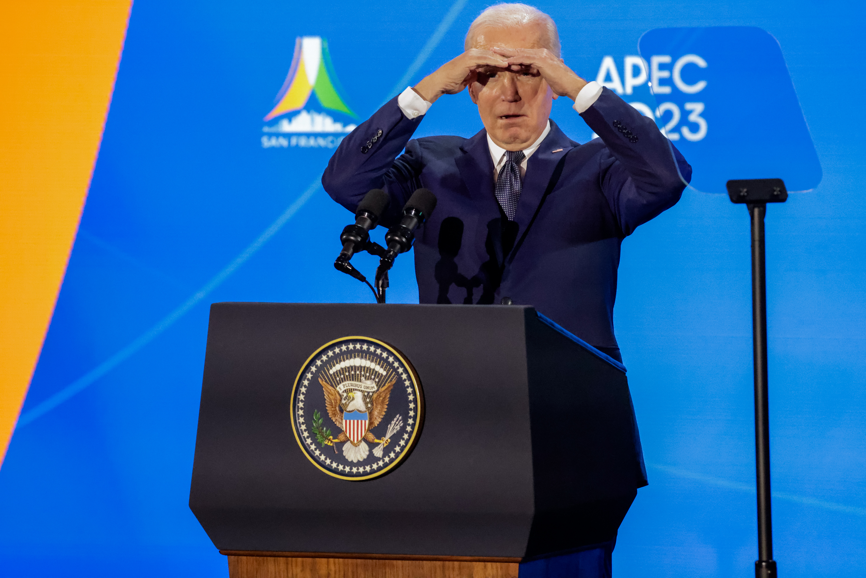 President Joe Biden holds his hands above his eyes to block a glare while standing behind a podium with APEC 2023 in the background.