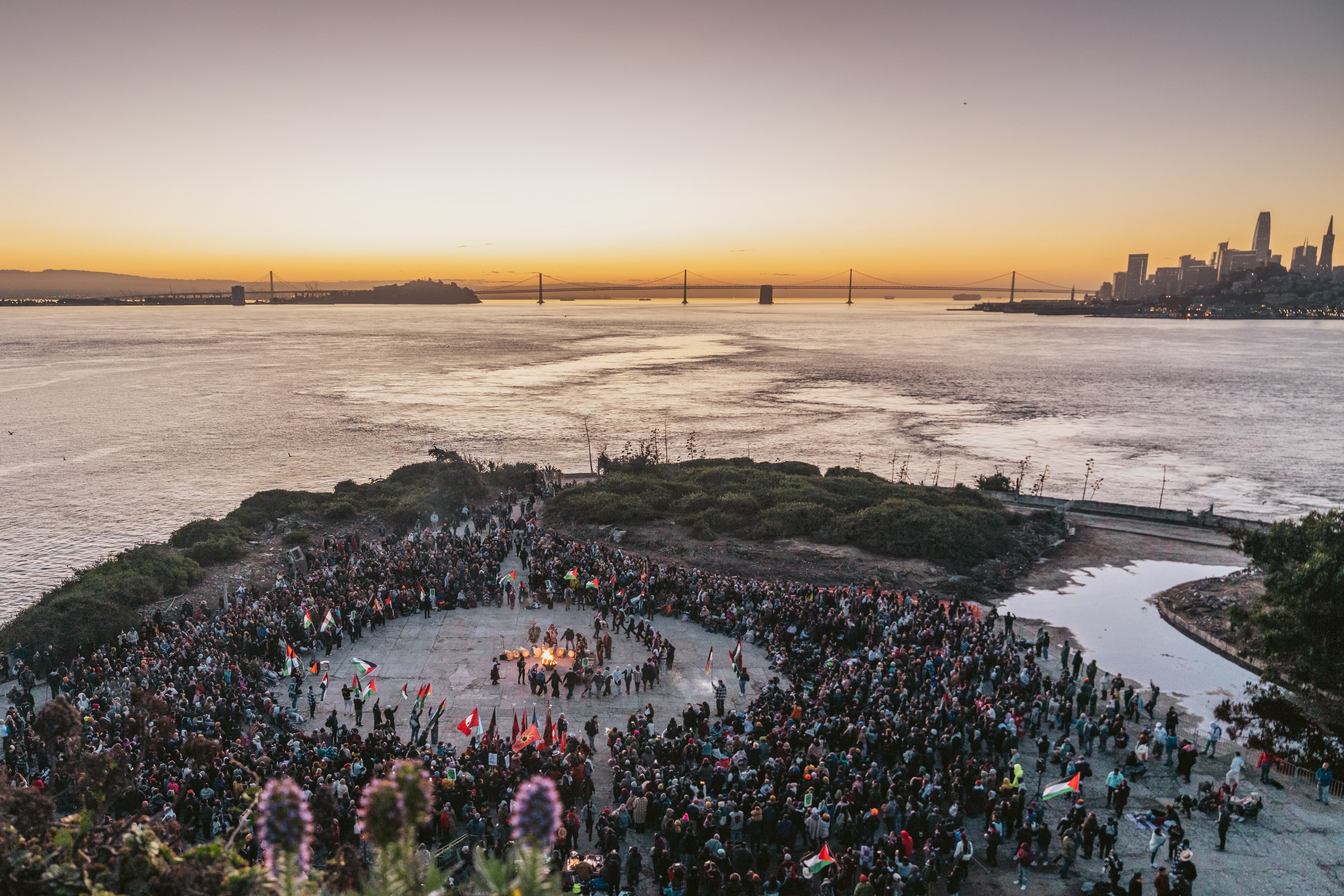 An overhead view of a crowd watching ceremonial dancing on an island with a view of San Francisco across the bay.