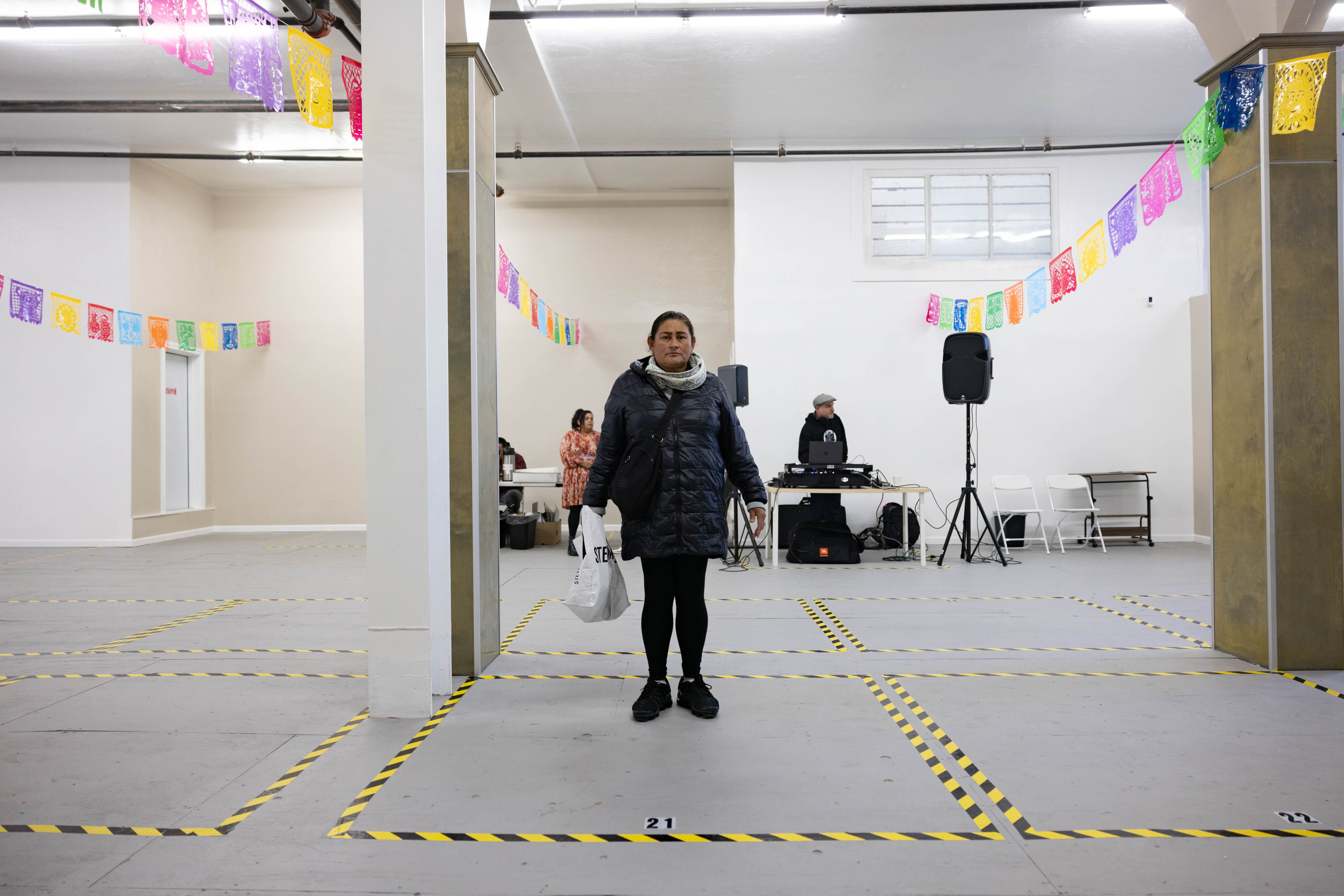 A women stands on the center of a taped off black and yellow rectangle in an empty warehouse looking space with a lone DJ in the background.