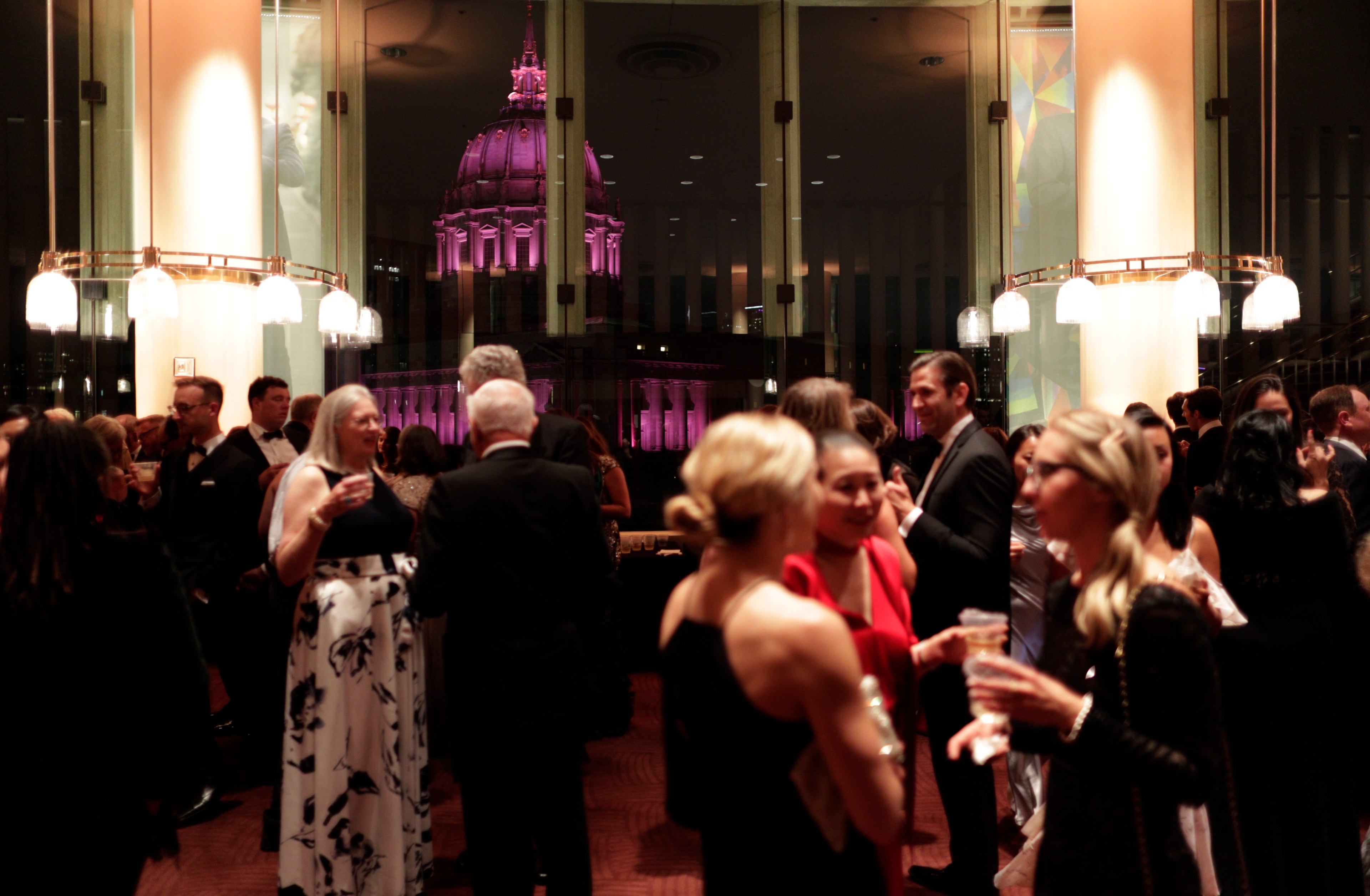 Dozens of people stand around talking during a cocktail hour in a lowly lit lounge space with a view of San Francisco City Hall out the window lit up in purple light.
