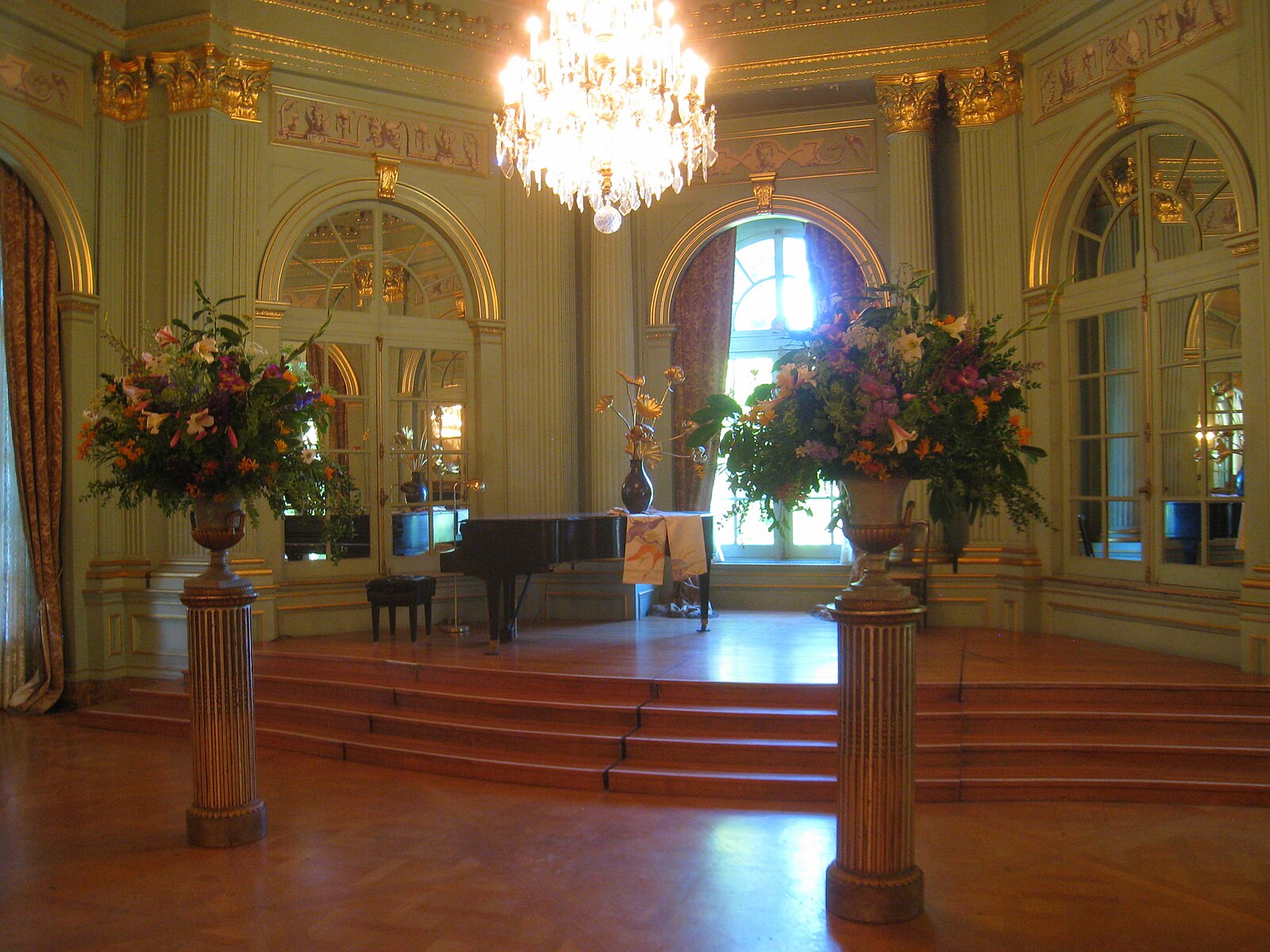 A piano atop a small stair case in an Victorian era interior that includes large mirrors and two large bouquets of flowers on pedestal, and a chandelier in the center of the room.