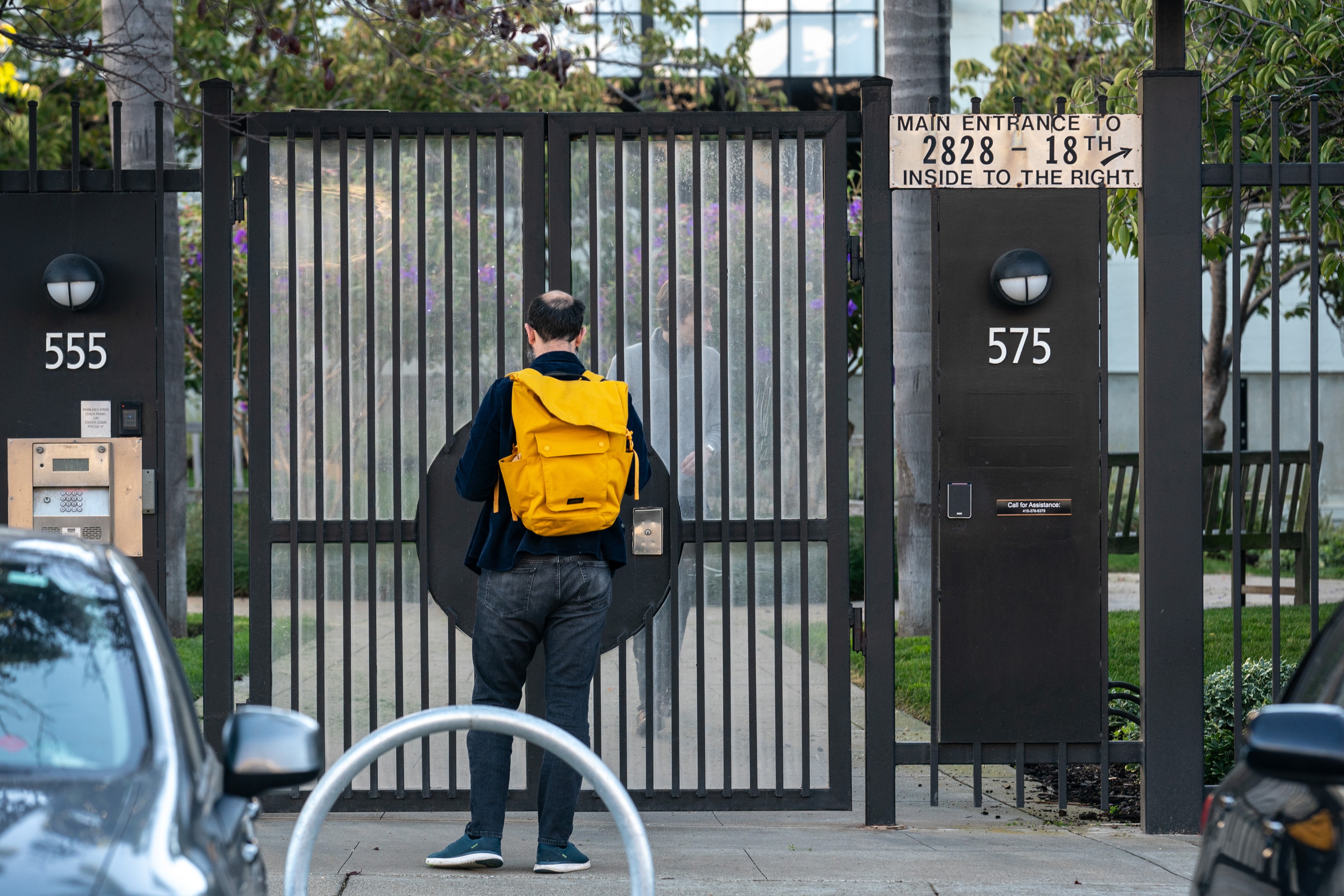 A person wearing a yellow backpack enters a large gated entrance with the street numbers 555 and 575 on the posts