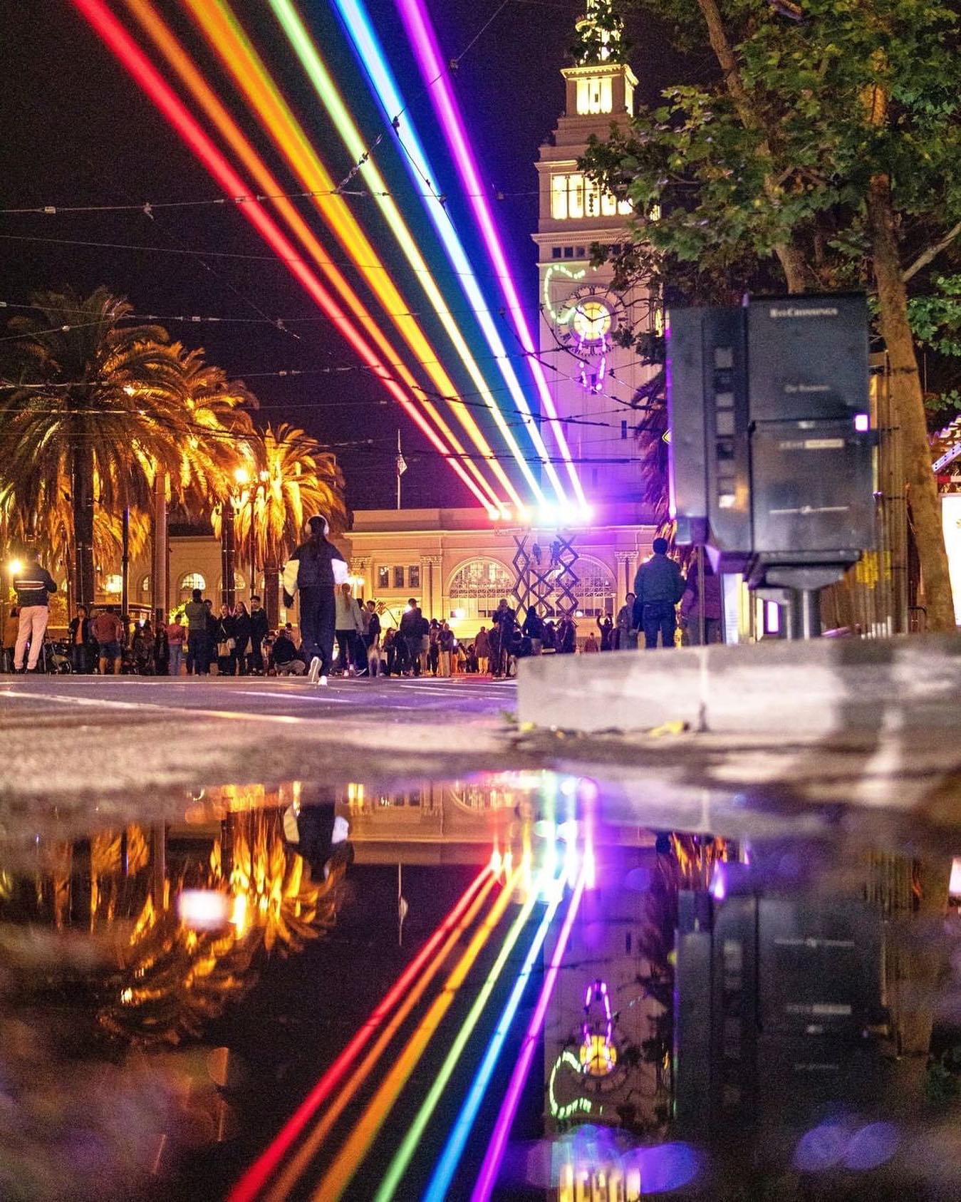 Rainbow laser beams shining over Downtown San Francisco are reflected in a puddle on the street below.