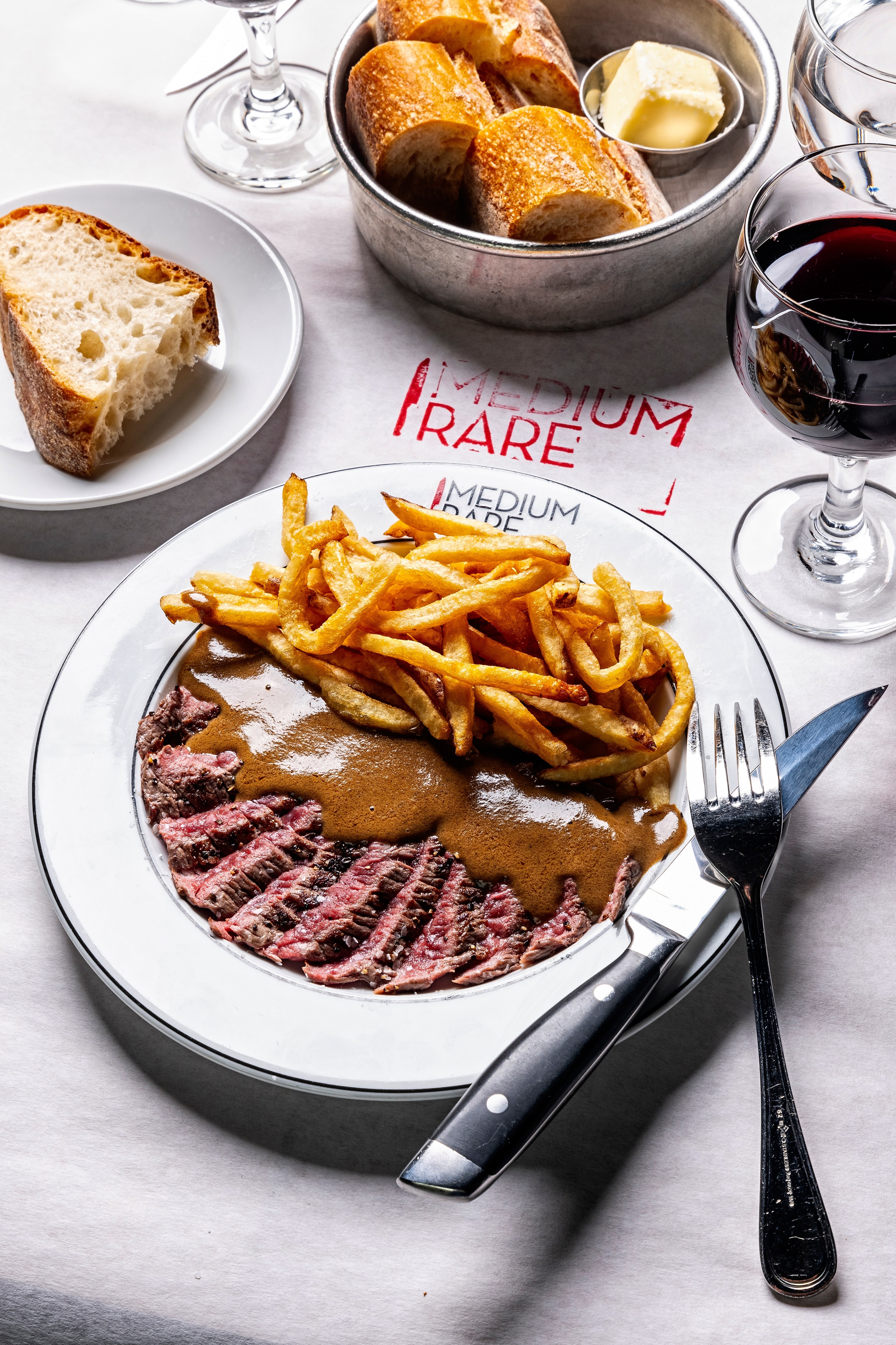 Two plates hold a large thick cut slice of fresh bread and steak, fries and sauce, joined by a glass of red wine and a metal bowl with more bread and butter in a smaller metal bowl, atop a white tablecloth.